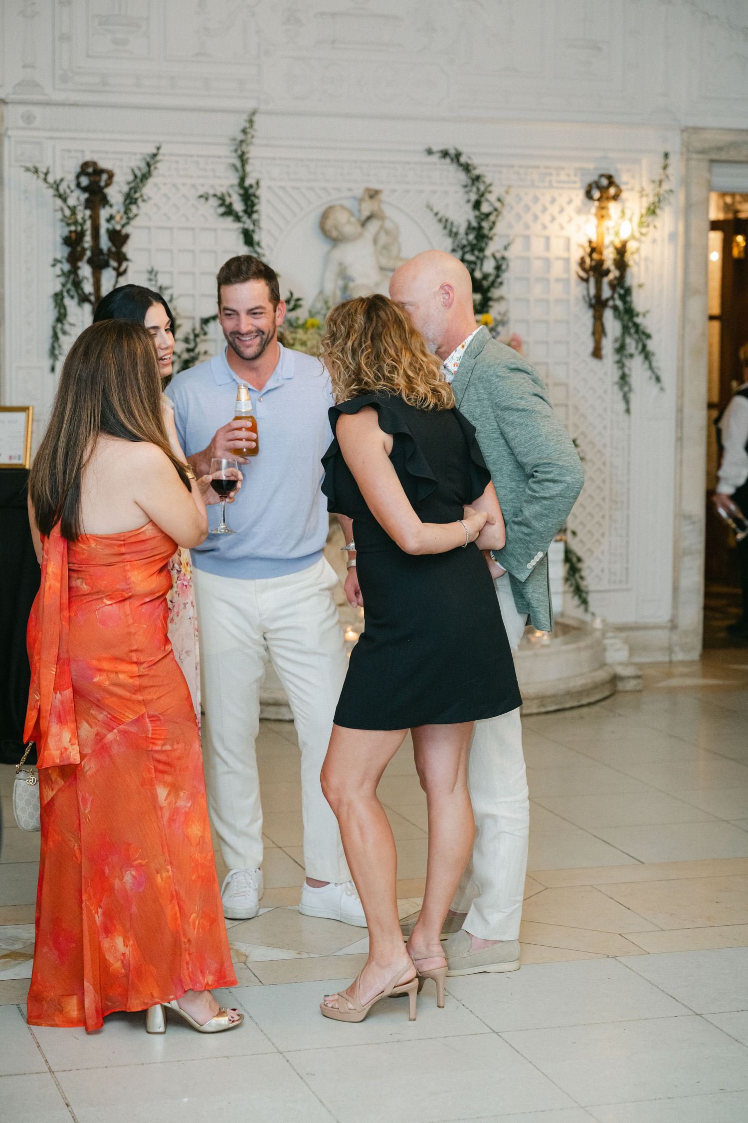 Two couples socializing at an elegant indoor event, woman in orange dress, man in blue polo holding wine glass.