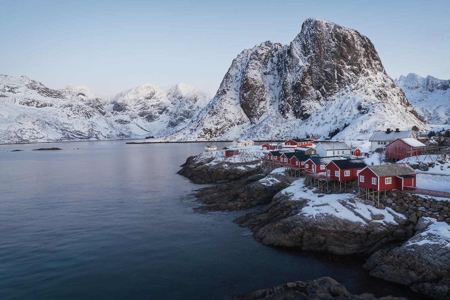 Red wooden cabins on rocky shore with snow-capped mountains reflected in calm Norwegian fjord waters.