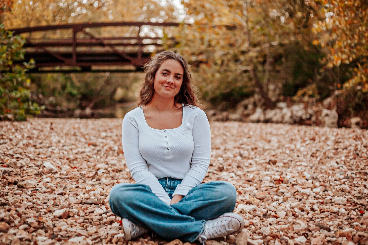 Young woman sitting cross-legged on autumn leaves near a wooden bridge, wearing jeans and white top.
