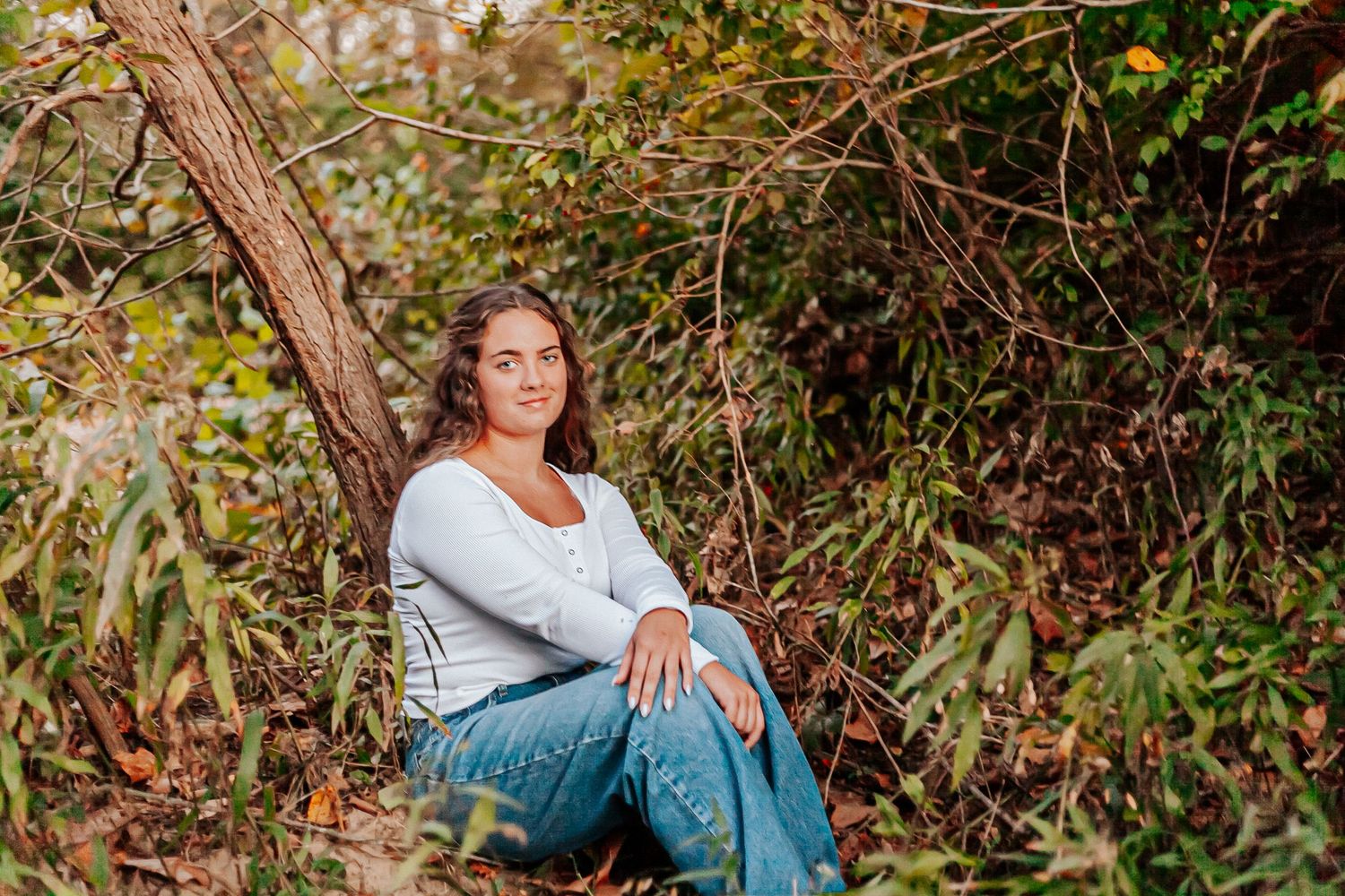 Woman in white top and jeans sitting peacefully against a tree surrounded by lush green foliage outdoors.