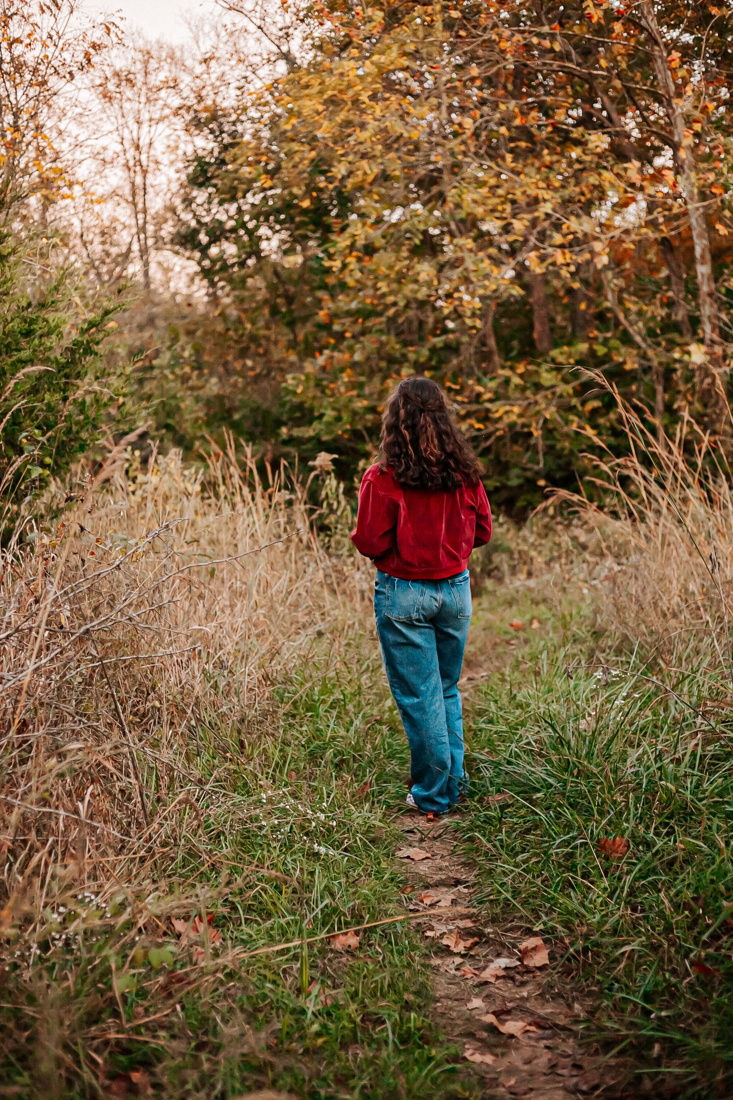 Woman in red top and jeans walking along a narrow autumn forest trail surrounded by tall grass and colorful foliage.