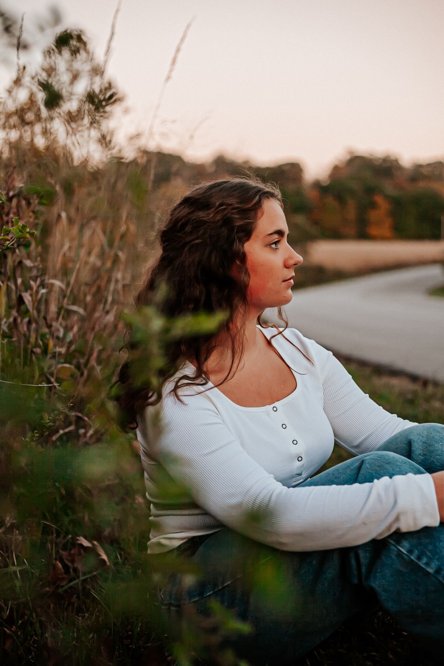Young woman in white top and jeans sitting outdoors in golden hour light, gazing thoughtfully into the distance.