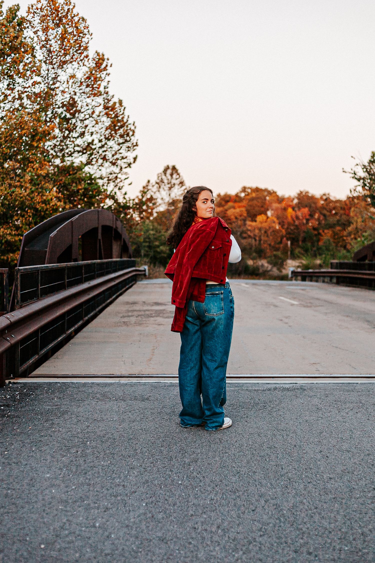 Young woman in red jacket and wide-leg jeans stands on a bridge surrounded by colorful autumn foliage at sunset.
