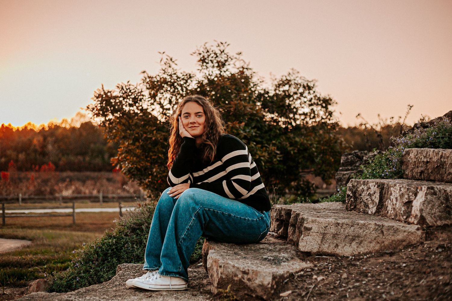 Young woman in striped sweater and jeans sitting on stone steps at sunset with autumn foliage background.