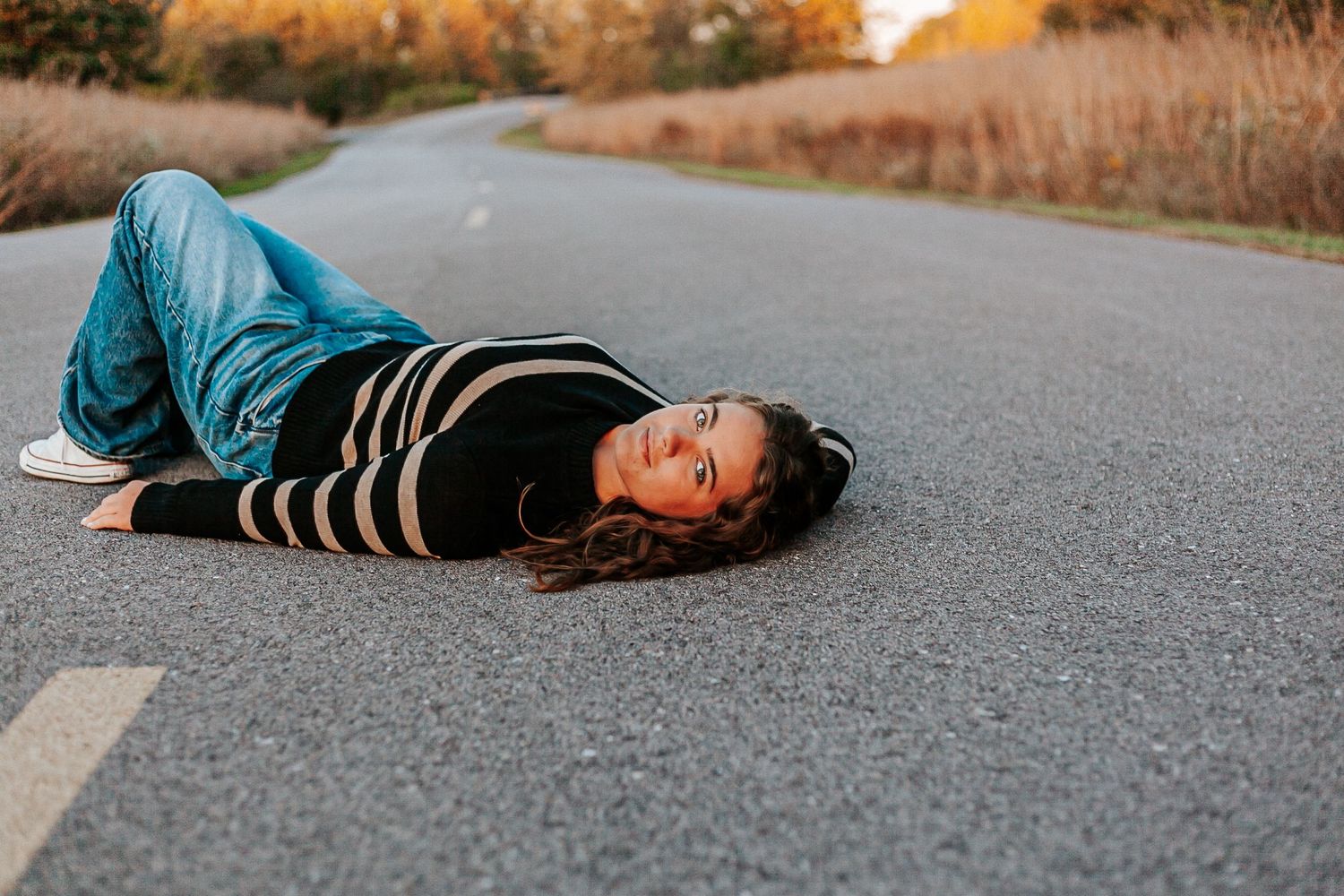 Young woman lying on a winding road wearing blue jeans and striped black sweater at sunset.