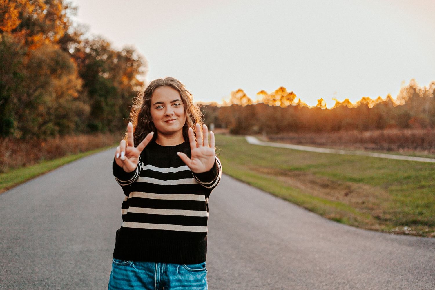 Teen girl in striped sweater standing on a road showing seven fingers, surrounded by autumn trees at sunset.