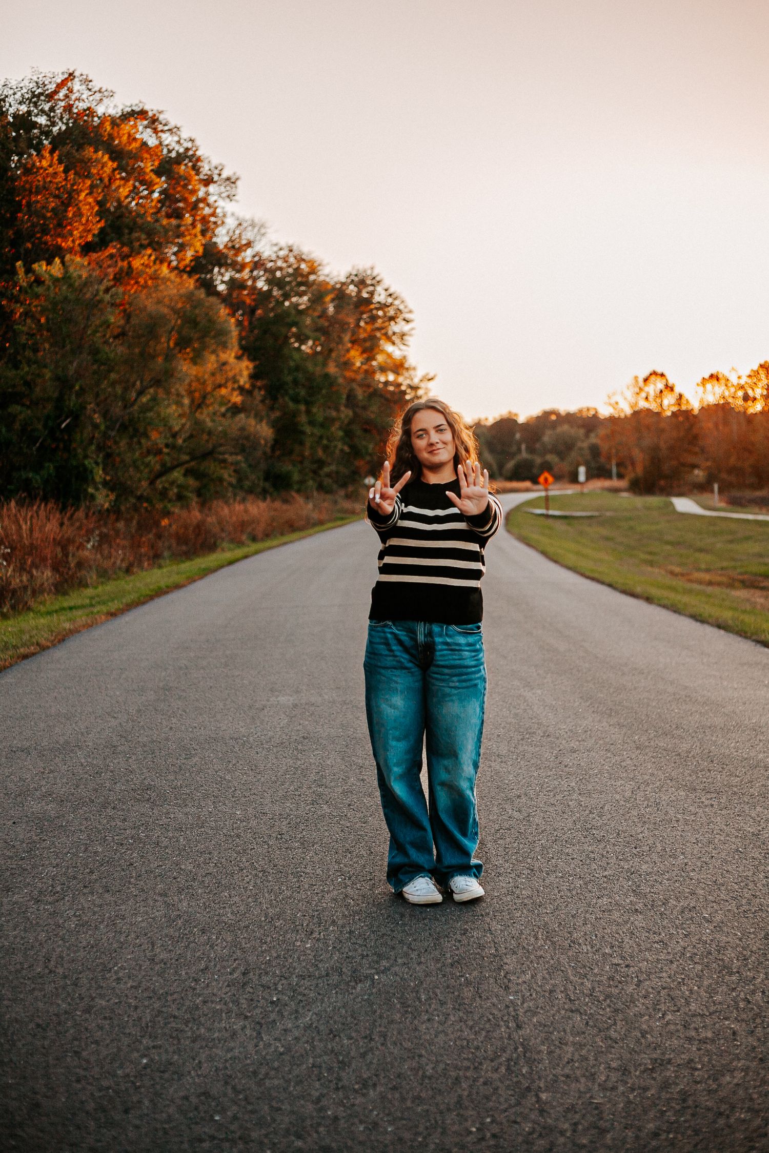 Young woman in striped sweater and jeans making peace signs on a scenic road surrounded by autumn foliage at sunset.