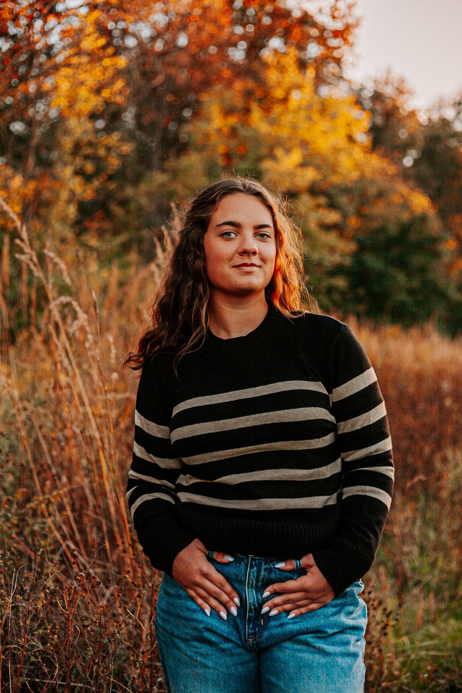 Young woman in striped sweater and jeans poses in autumn field with colorful fall foliage backdrop.