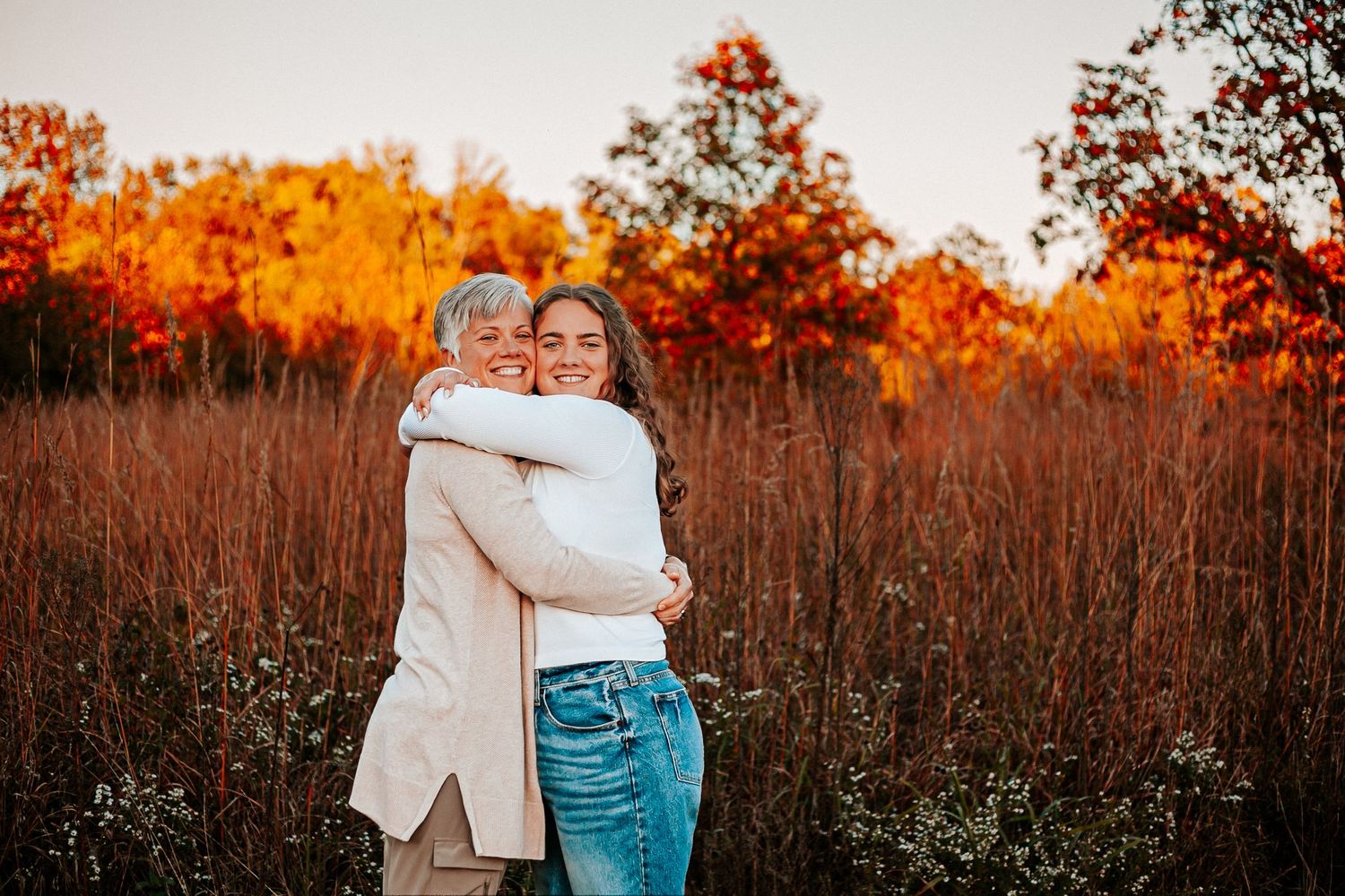 Two women embrace in a field of tall grass with vibrant autumn foliage in shades of red and orange behind them.