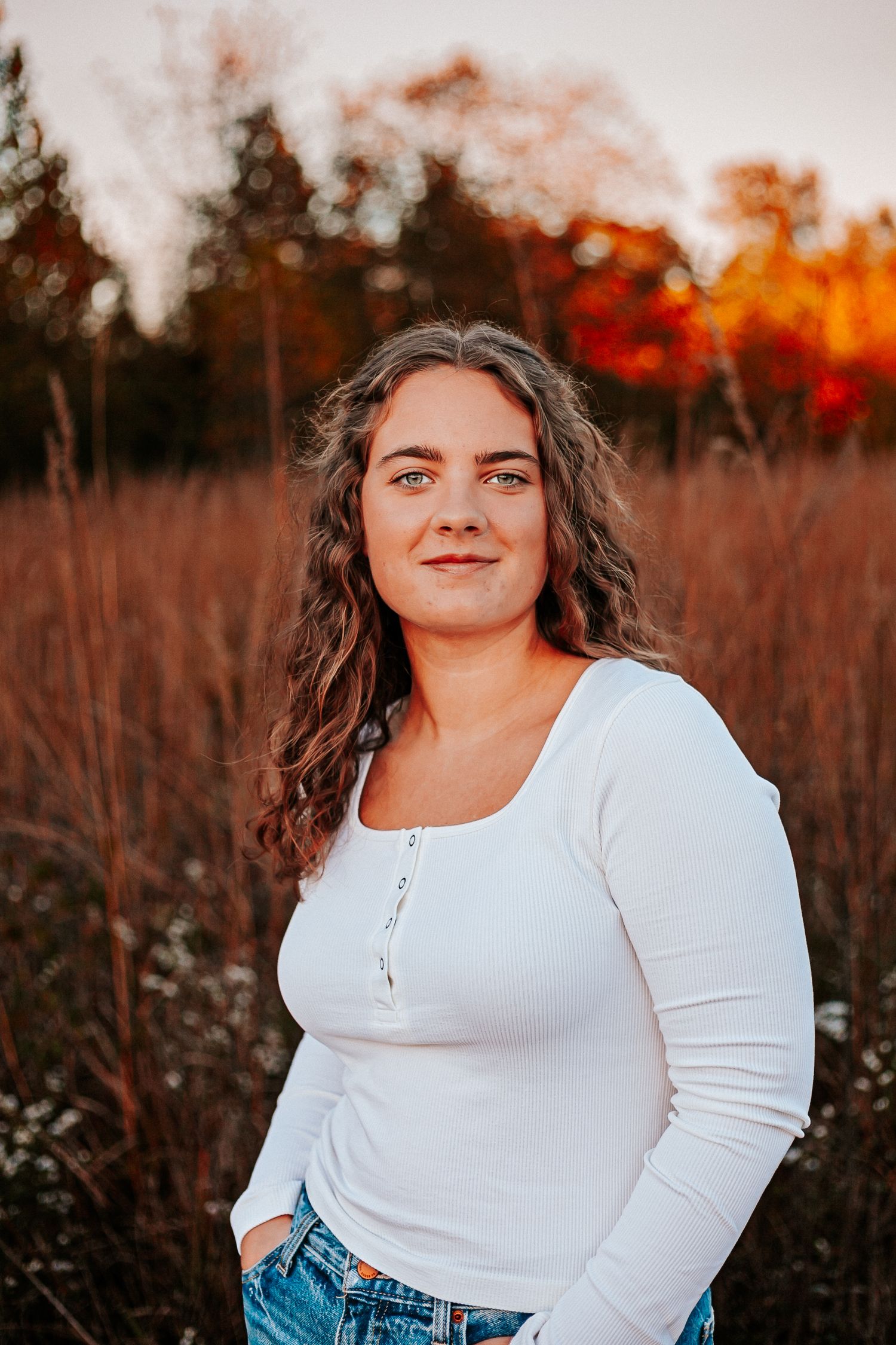 Young woman in white long-sleeve top and jeans stands in golden autumn meadow at sunset.