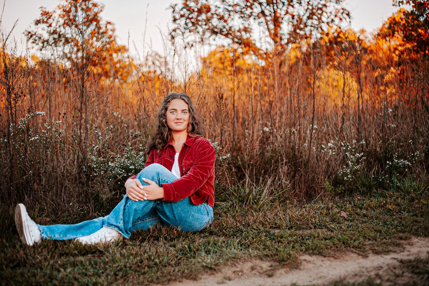 A young woman in a red jacket and jeans sits on grass near a dirt path surrounded by autumn foliage at golden hour.