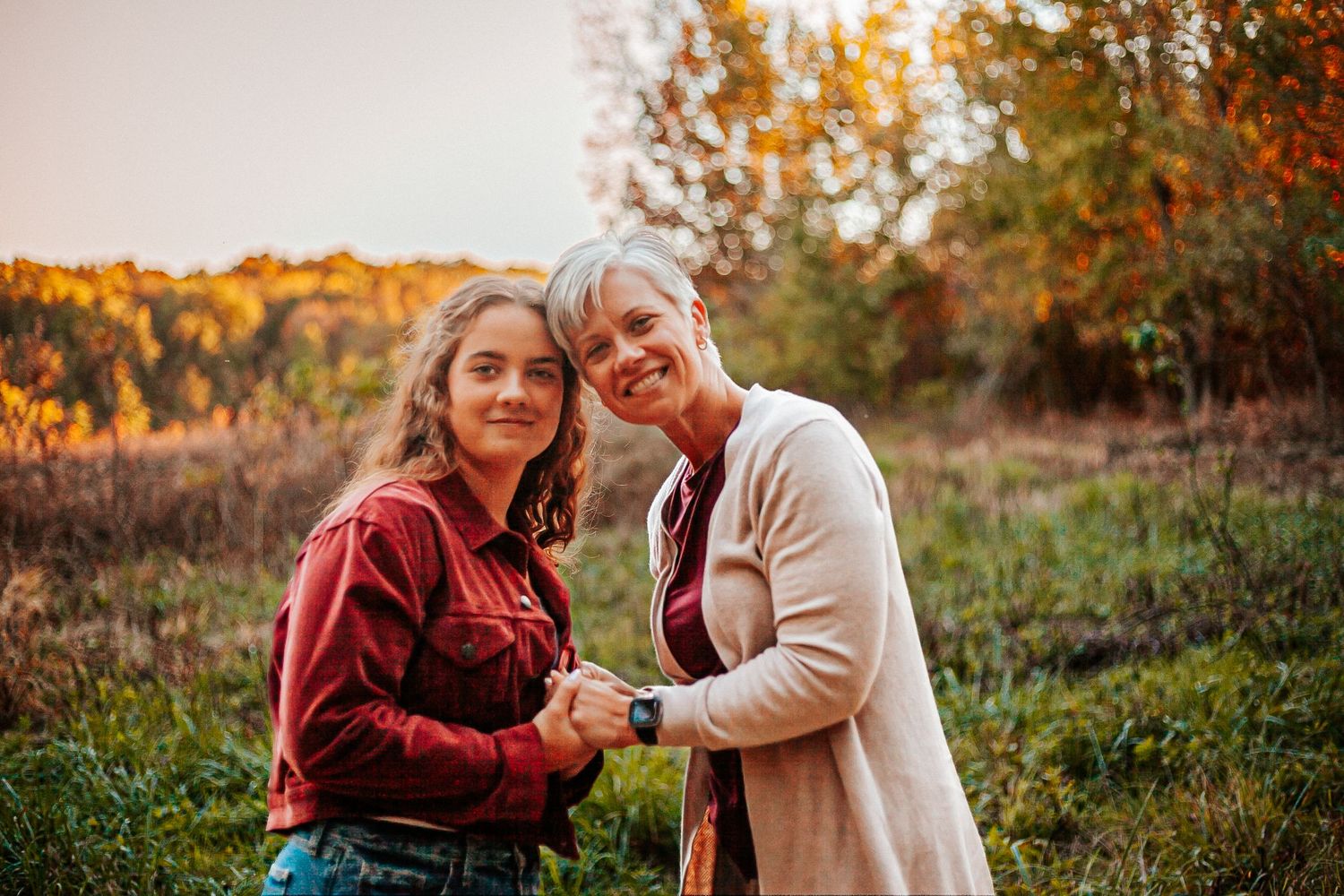 Two women smiling and holding hands in an autumn field with colorful foliage at golden hour.