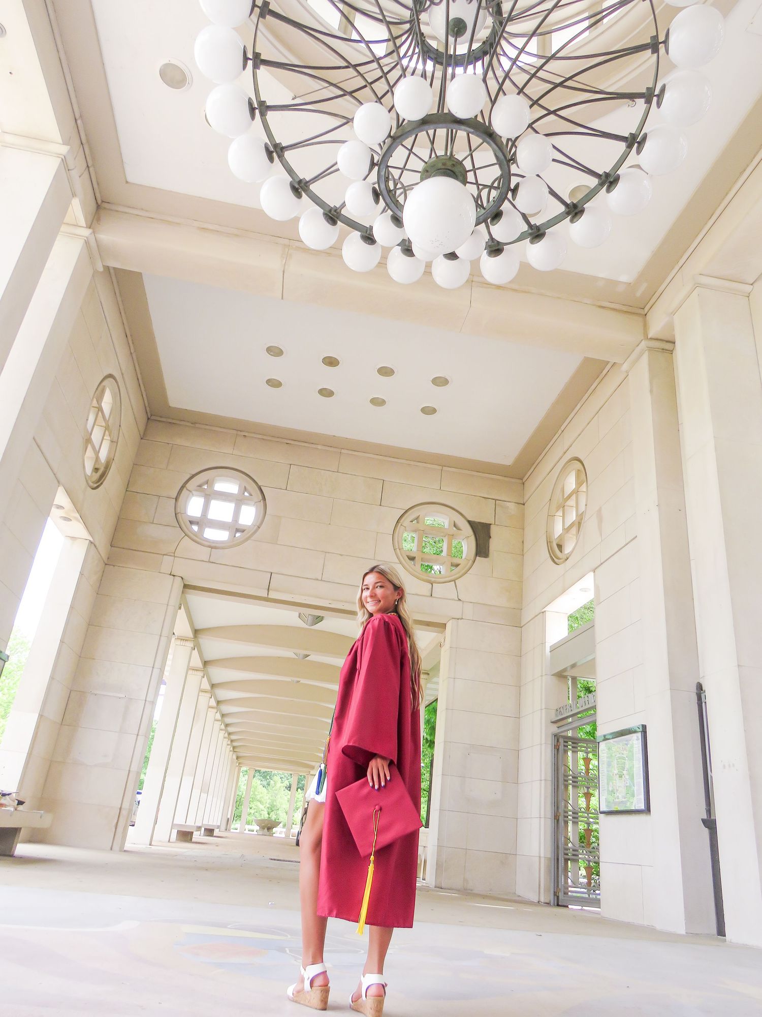 Woman in red graduation gown and cap posing under elegant chandelier in a grand columned hallway.