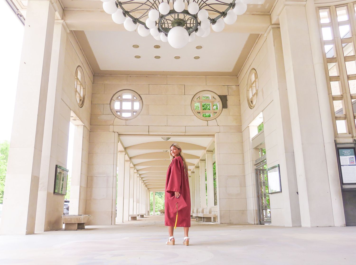 Woman in red coat and pants stands in elegant columned arcade with ornate chandelier and arched windows.