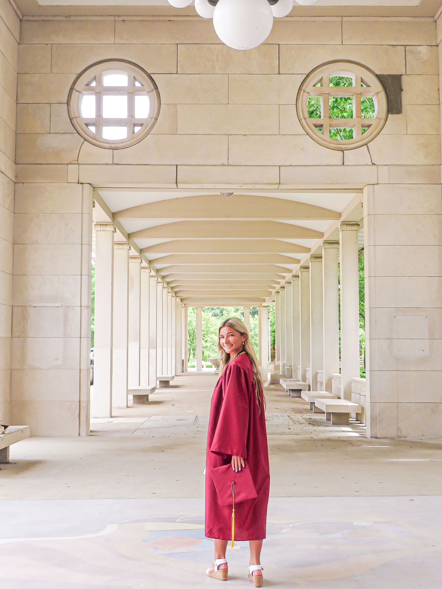 A smiling graduate in a crimson cap and gown poses in an elegant stone colonnade with arched ceilings and circular windows.