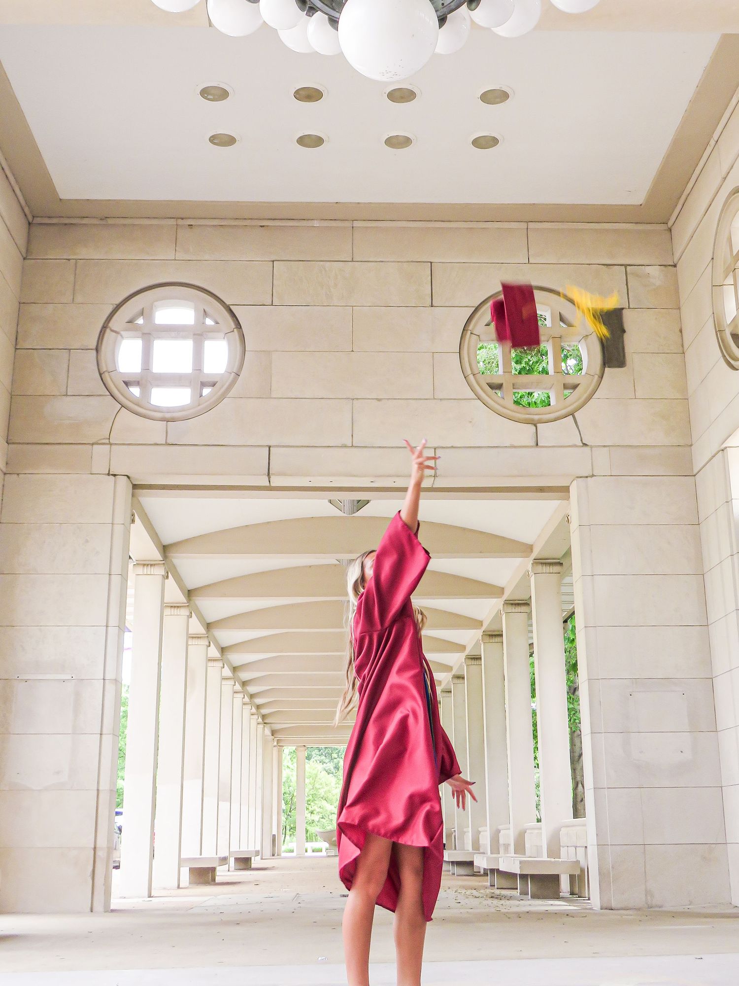 A graduate in a red gown raises her hand in a elegant stone colonnade hallway.