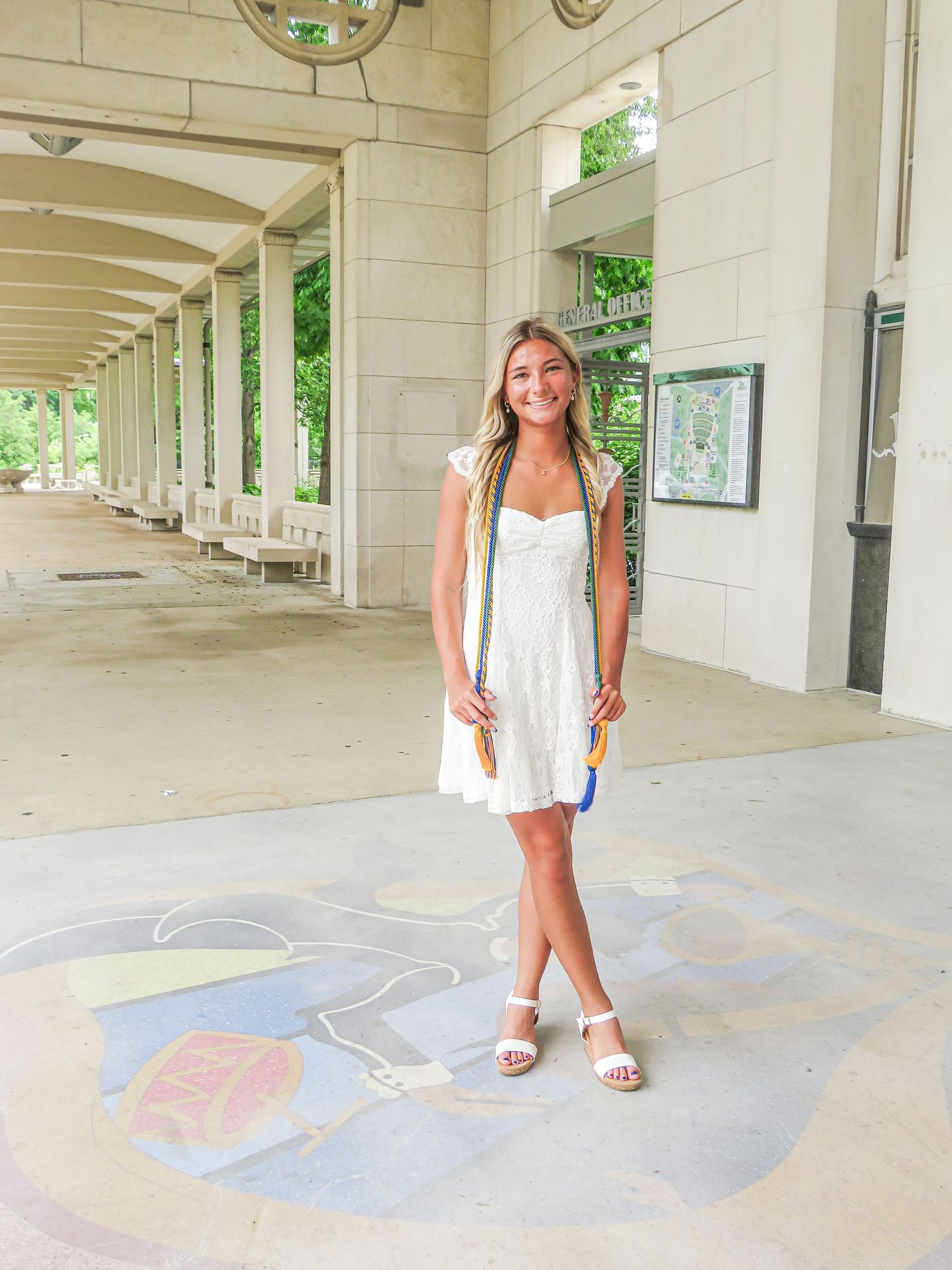 Smiling young woman in white mini dress and sandals standing under a covered colonnade walkway.