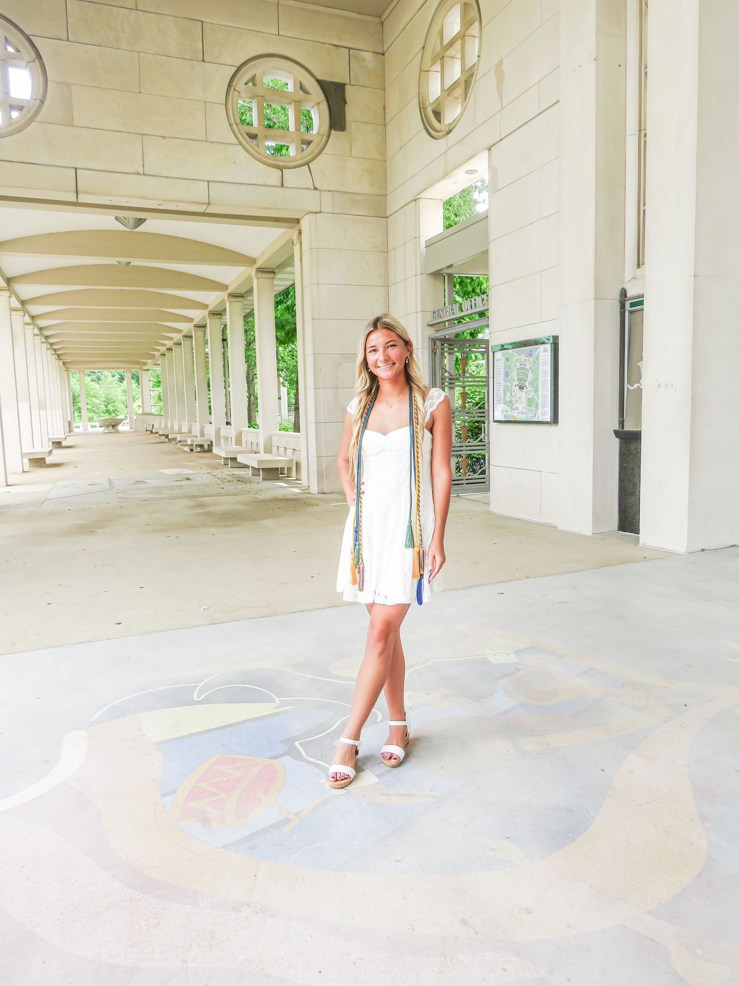Young woman in white dress and sandals holding graduation tassel, standing under arched colonnade on decorated pavement.
