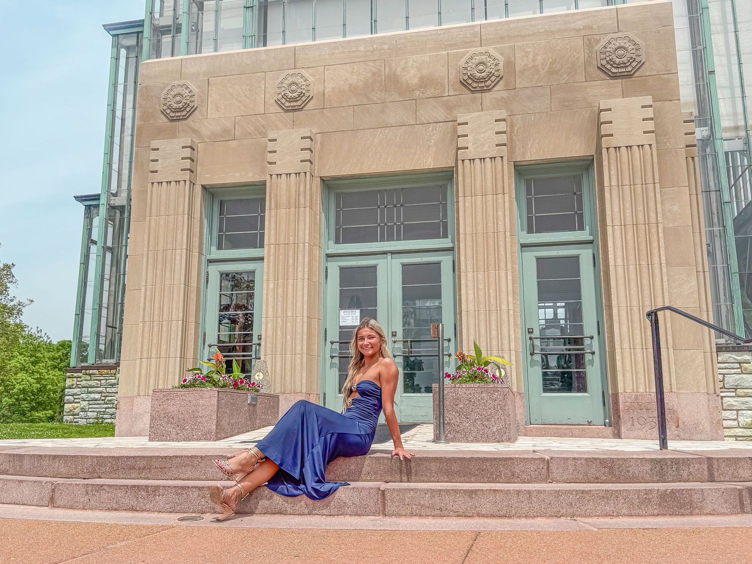 Woman in blue gown posing on steps of elegant stone building with decorative columns and teal doors.