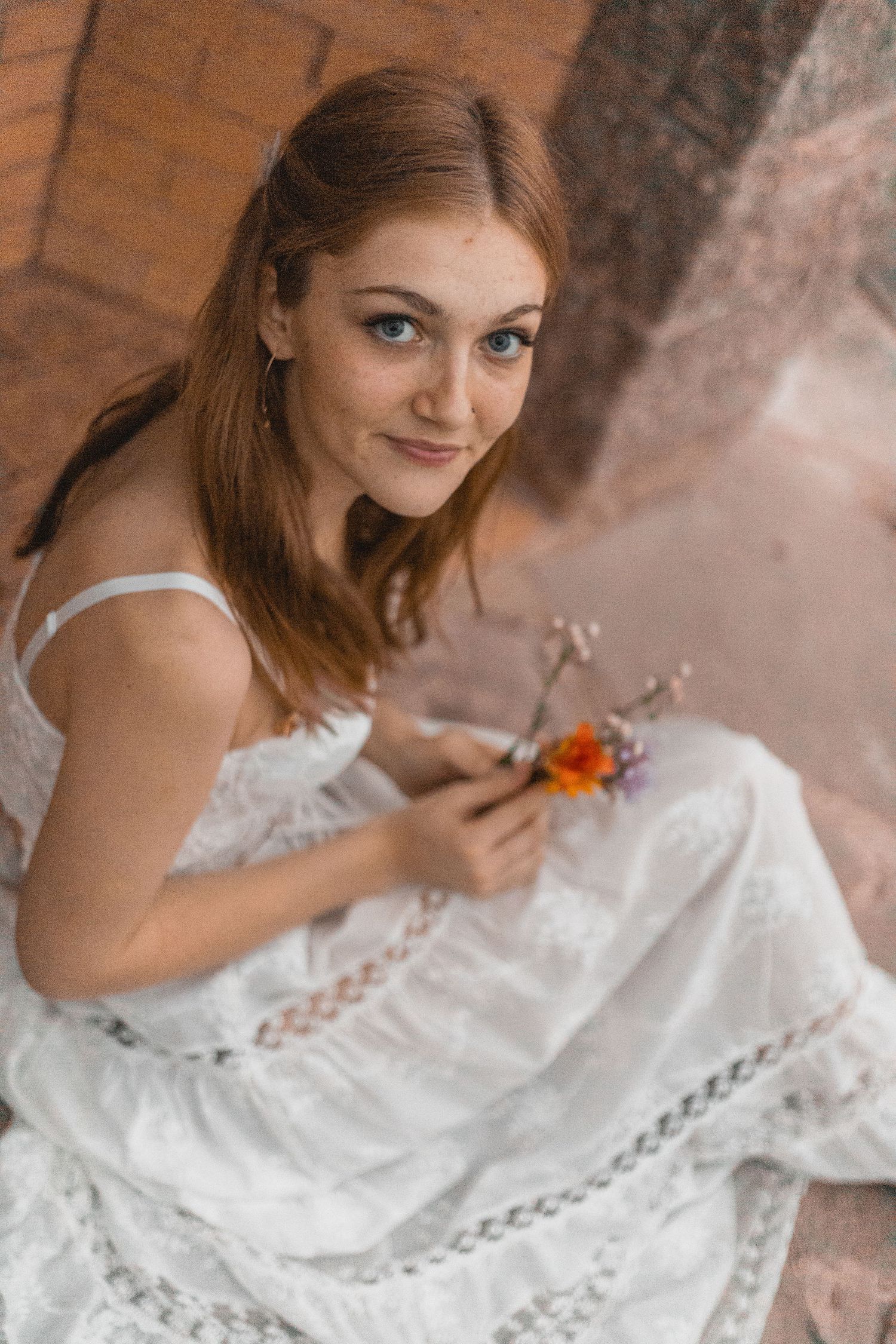 Young woman in white dress sitting against brick wall holding small wildflowers, smiling at camera.