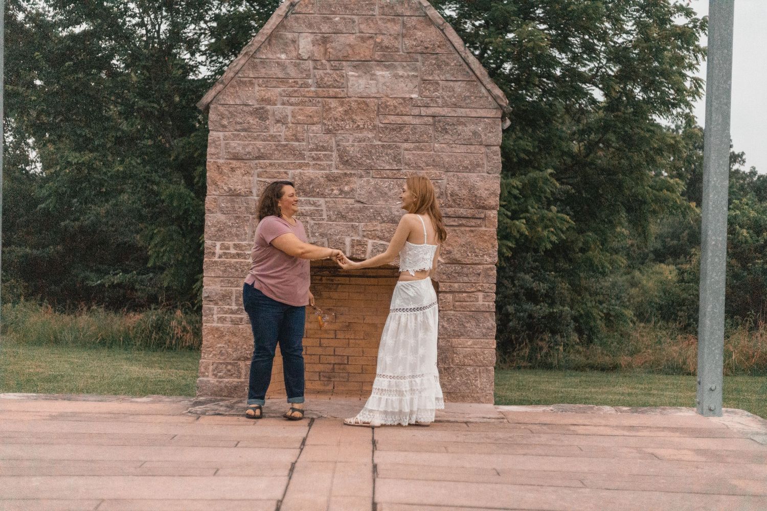 Two women stand at a rustic brick outdoor altar in a lush green park setting, sharing a meaningful moment.