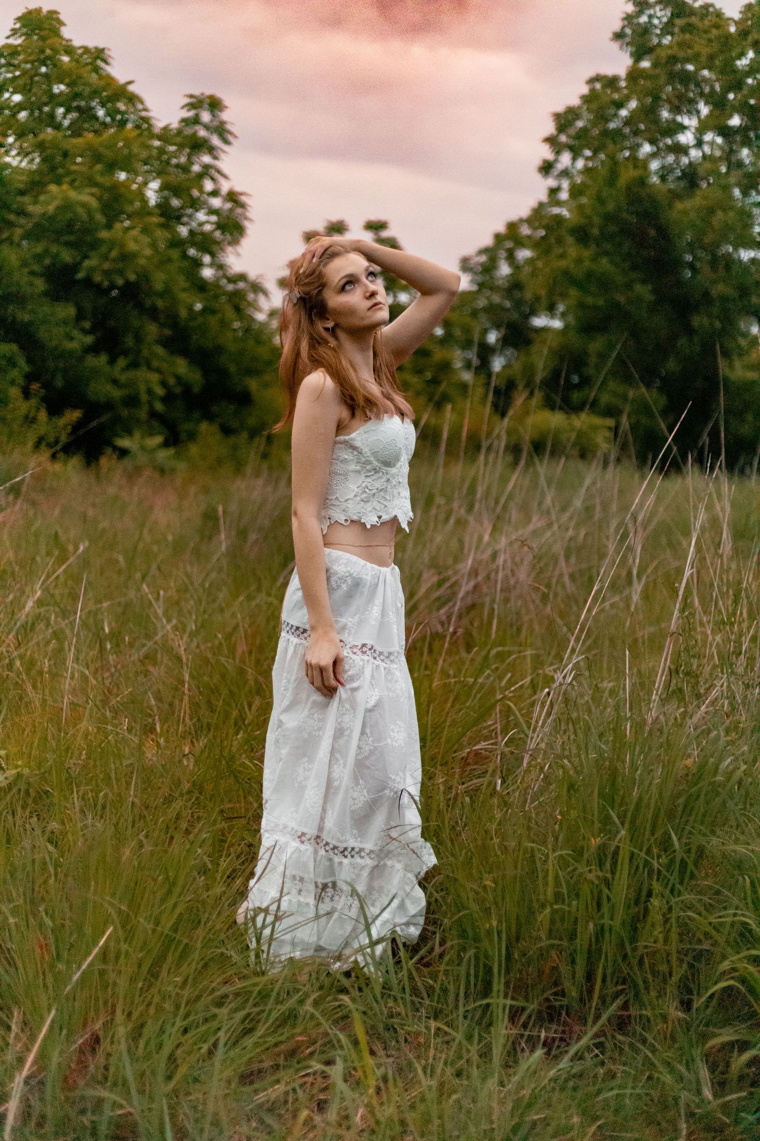 Young woman in white boho lace crop top and maxi skirt standing in tall grass field at sunset.