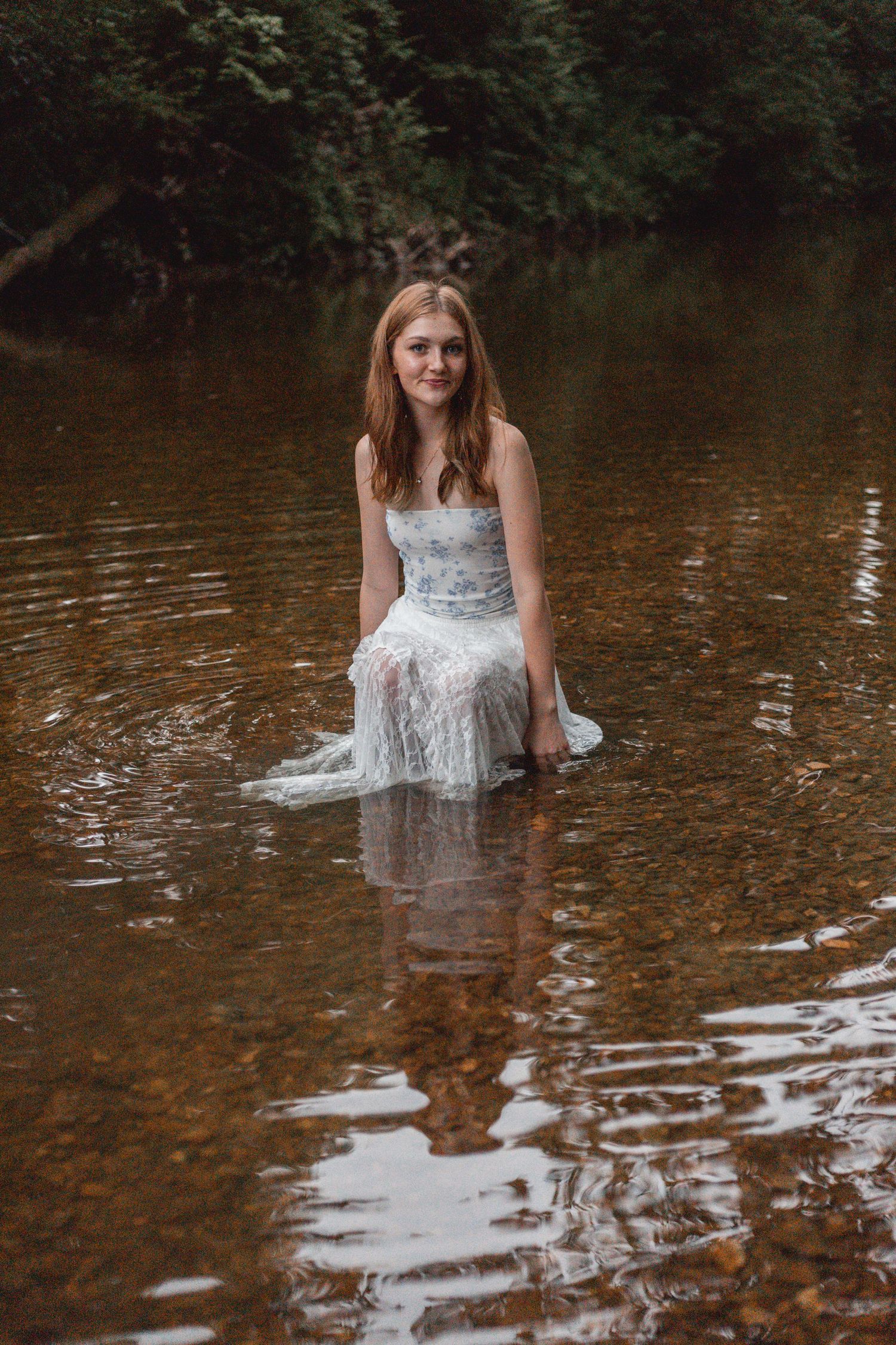 Young woman in white lace dress sitting in a shallow forest river, smiling at camera.