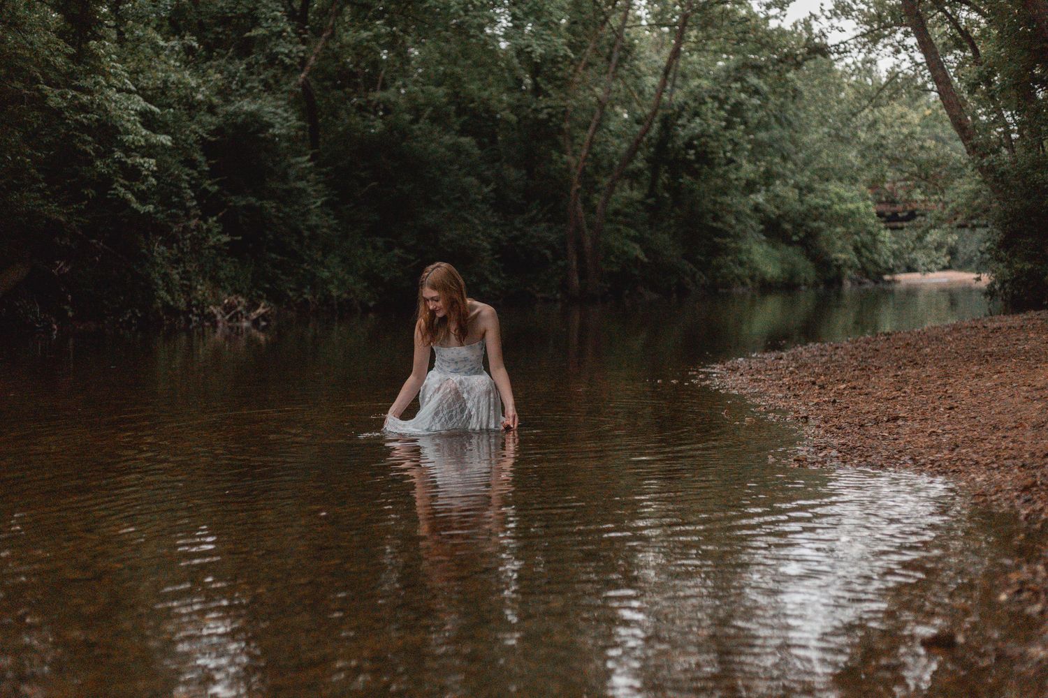 Woman in white dress wading through a calm forest river surrounded by lush green trees.