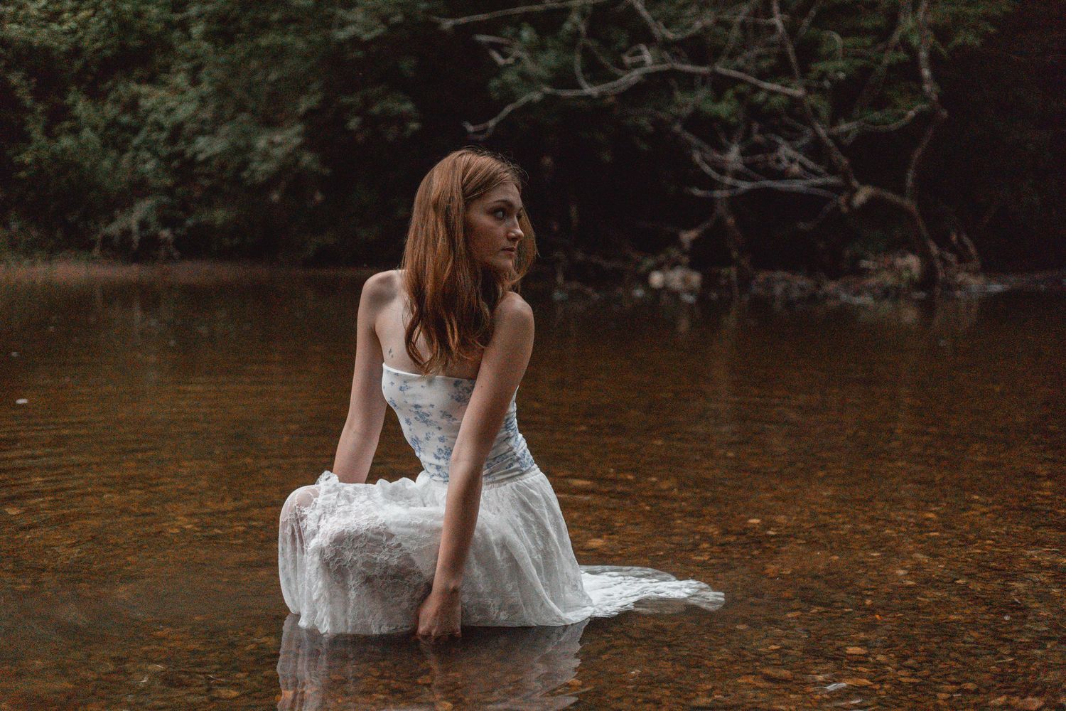 Red-haired woman in wet white lace dress sitting in shallow river with dark forest backdrop.