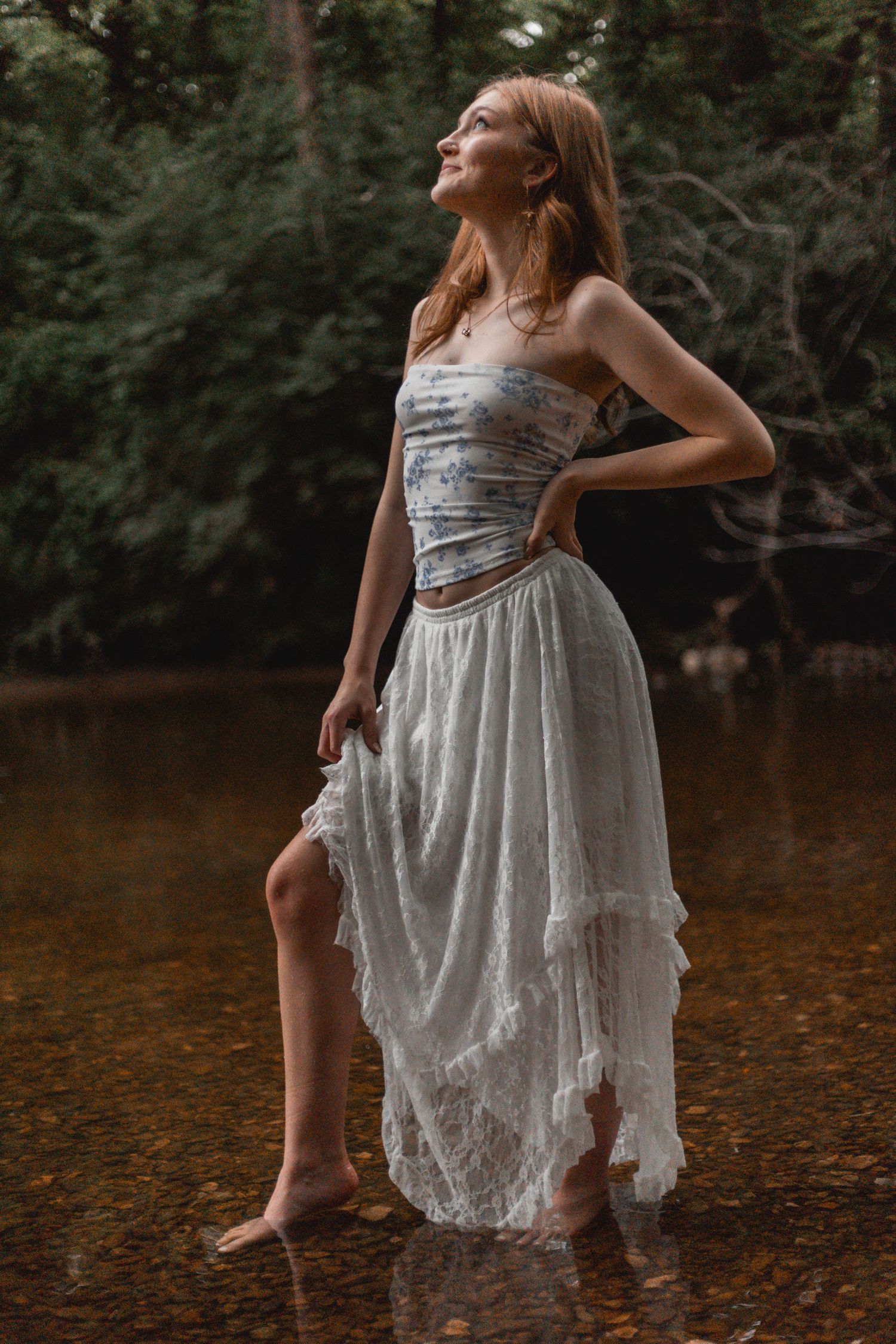 Woman in white lace skirt and blue corset top standing barefoot in a forest stream.