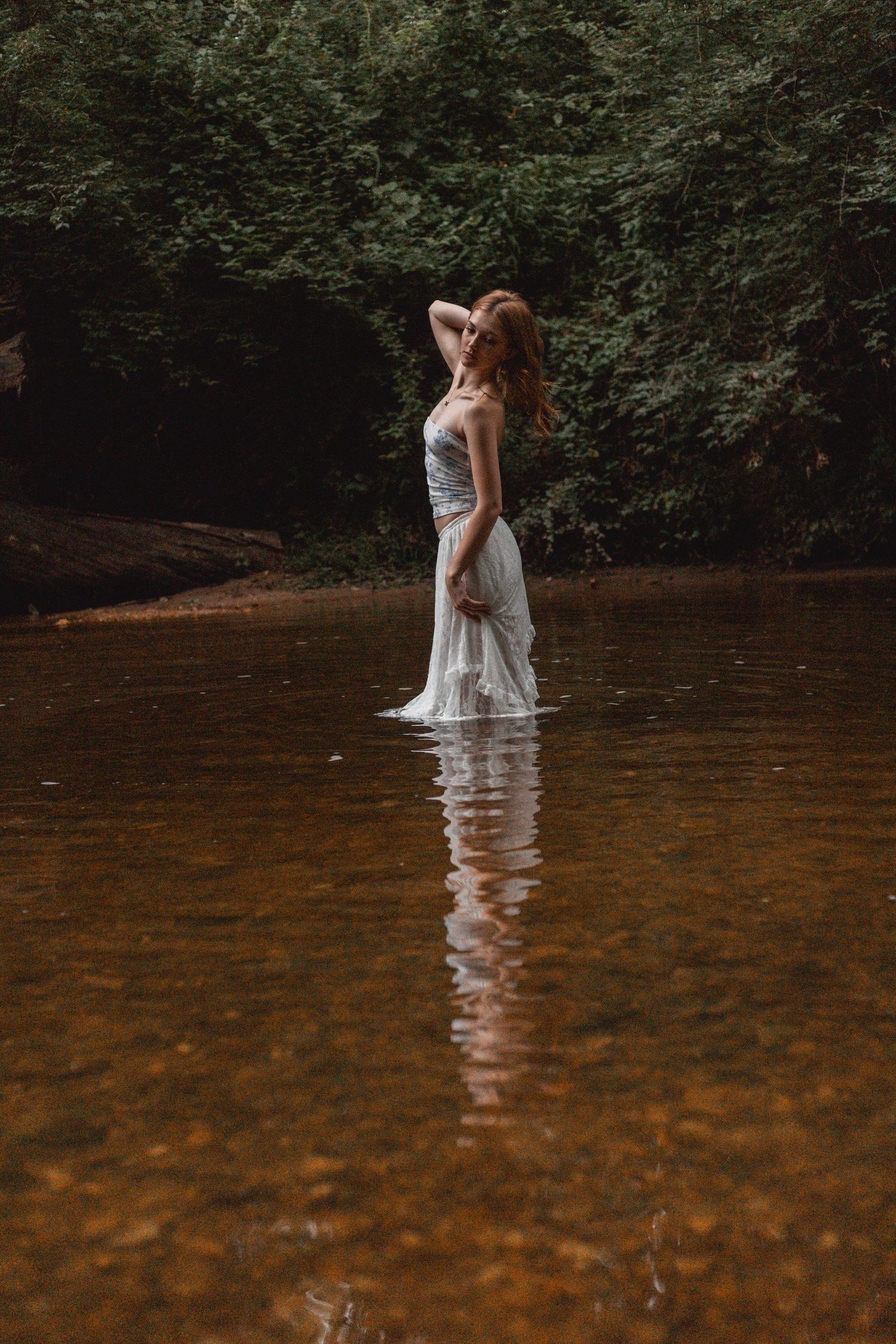 Woman in white dress standing in river with hands in hair, reflected in calm water.