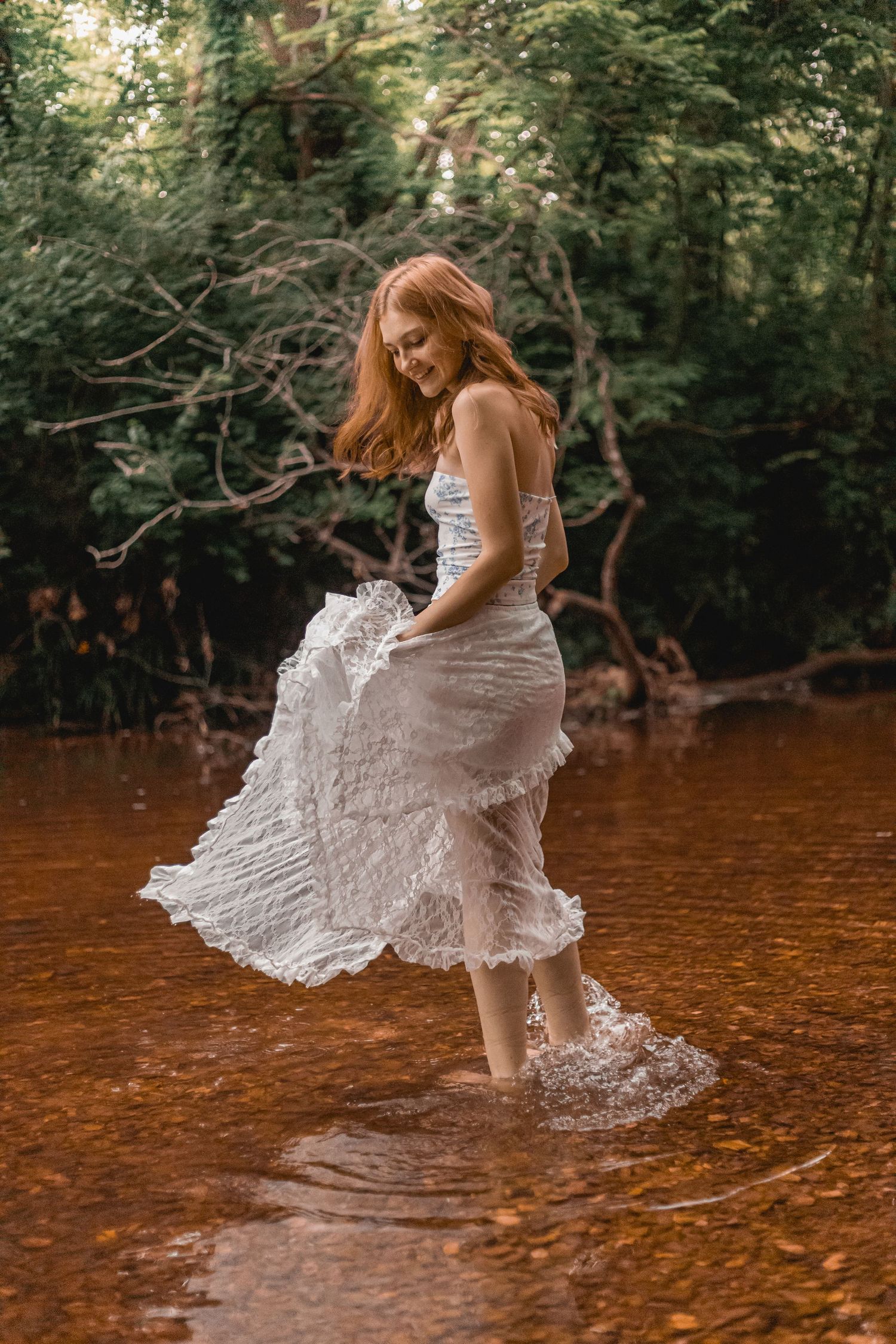Redhead woman in flowing white lace dress walking through shallow forest stream, laughing.