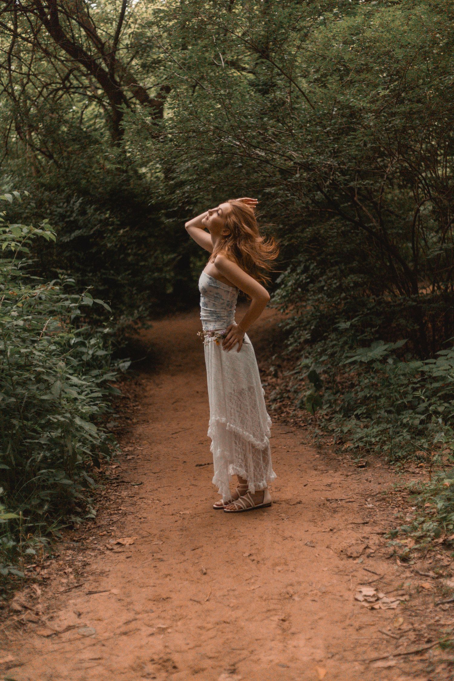 Woman in white lace dress posing on a forest dirt path surrounded by lush greenery.