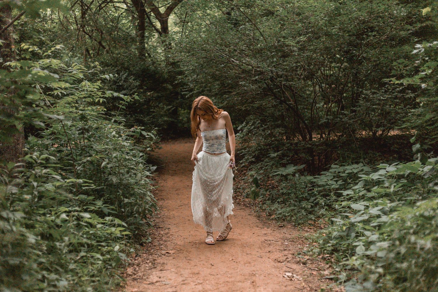 Young woman in flowing white dress walking along a lush forest path surrounded by green foliage.