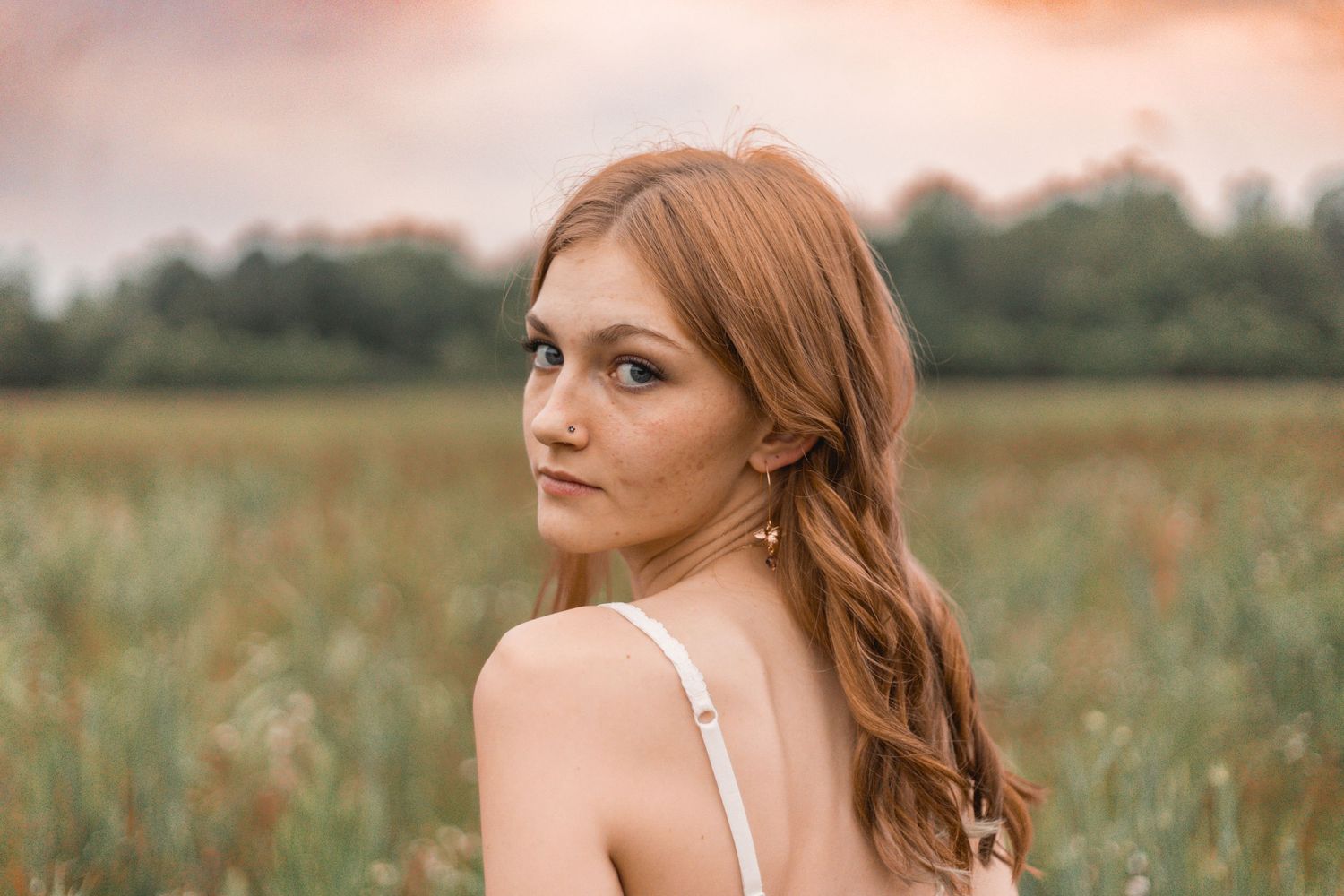 Beautiful redhead woman in white dress looking over shoulder in a field at golden hour.