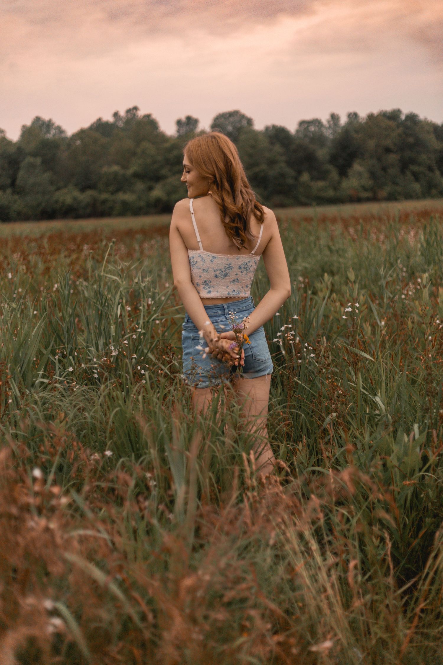 Young woman in floral crop top and denim shorts holding wildflowers in a green meadow at sunset.