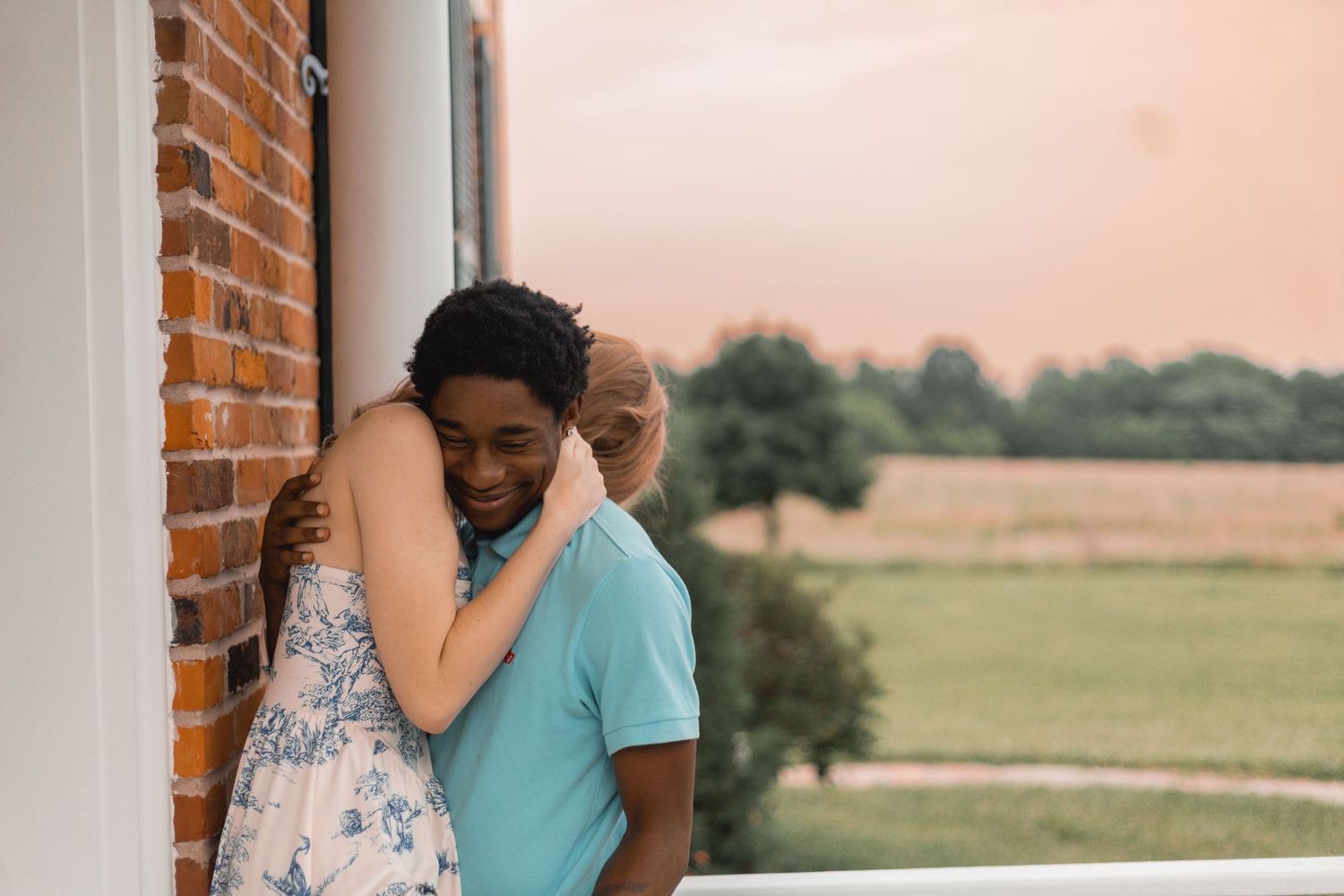 Happy couple embracing by brick wall on porch with scenic countryside sunset in background.