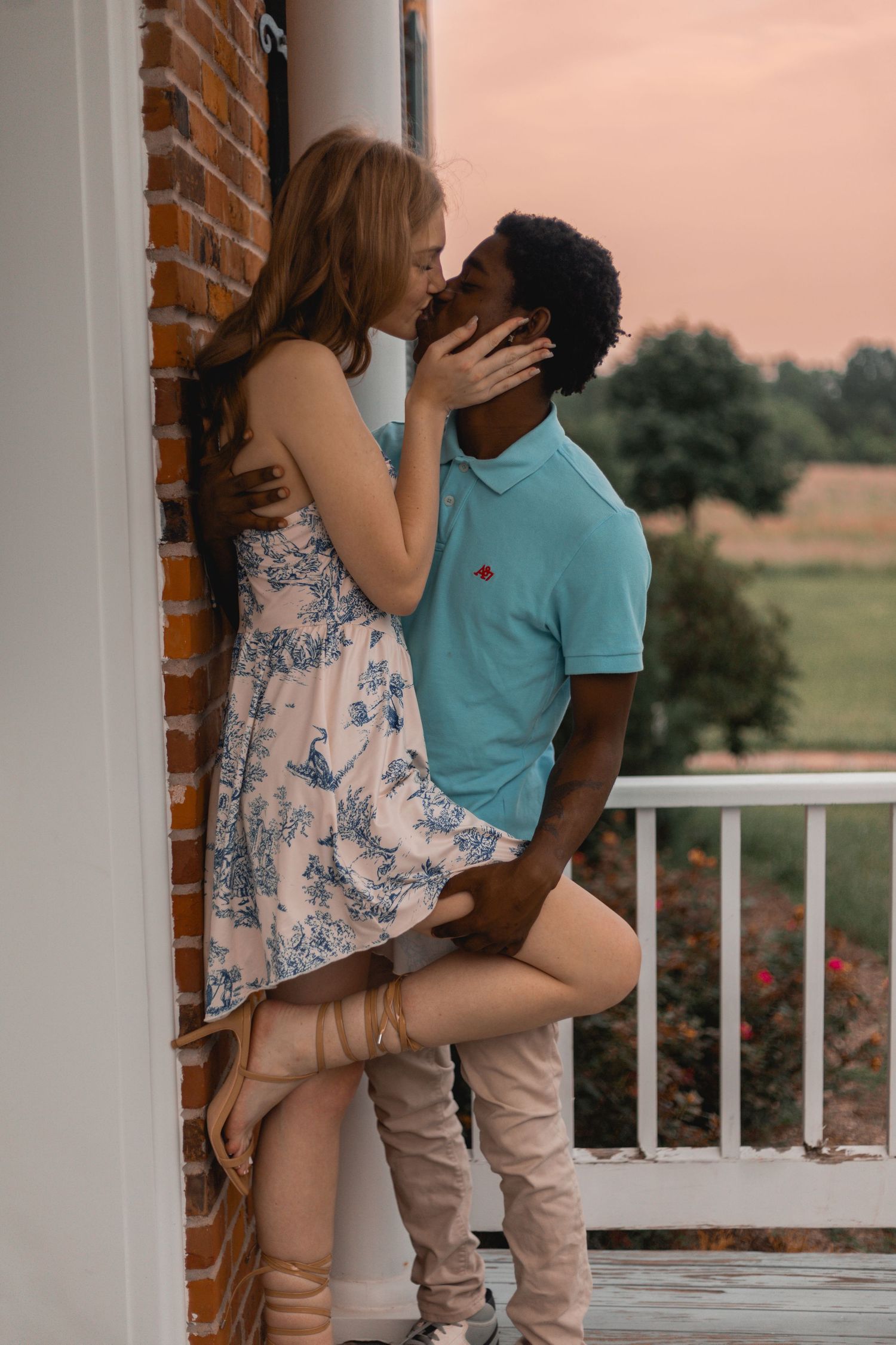 Couple sharing a kiss on a porch at sunset, woman in floral dress, man in teal polo shirt.