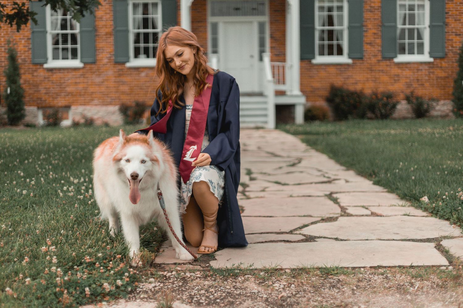 Graduate in cap and gown petting a white husky dog on stone path outside brick home.