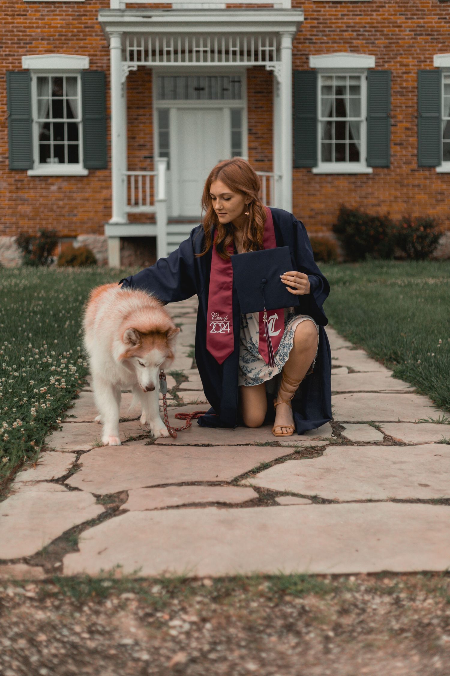 Young woman in graduation cap and gown kneeling beside a fluffy dog on a stone walkway.