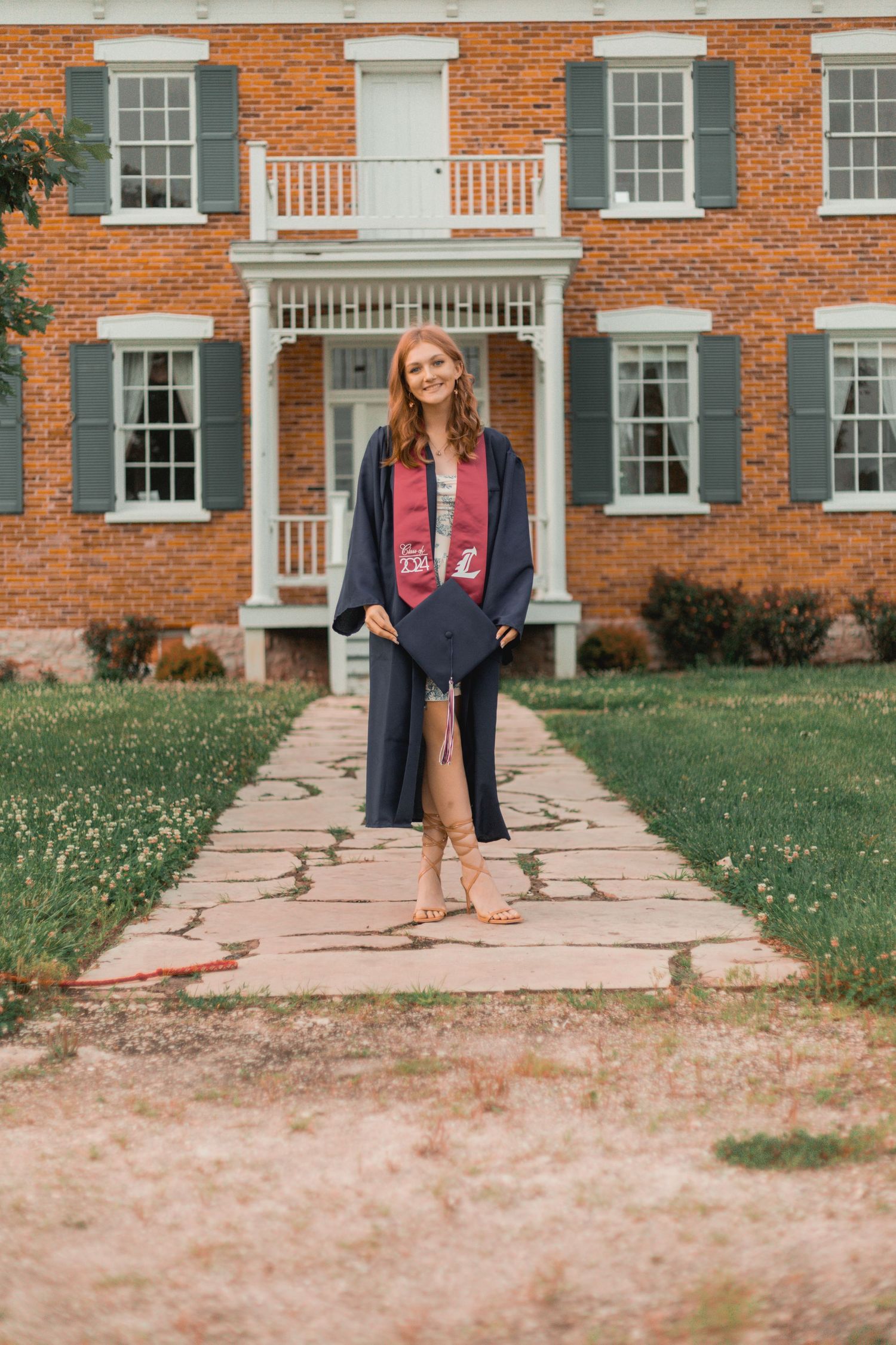 Smiling graduate in navy cap and gown with maroon stole posing outside brick building.