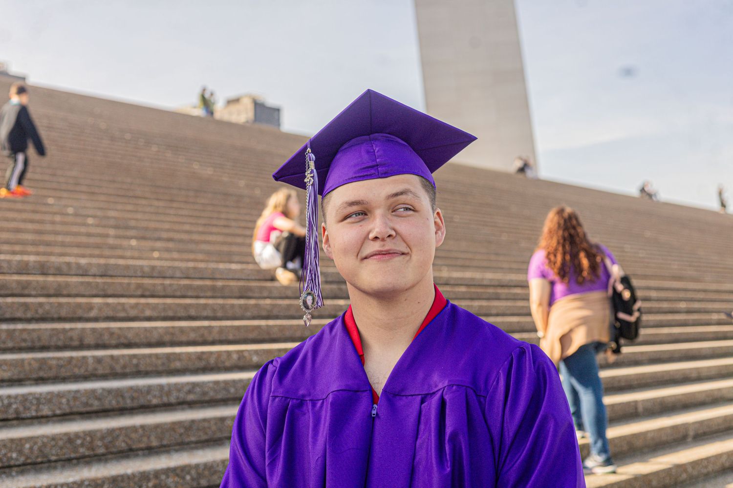 Young graduate in purple cap and gown smiling on monument steps at outdoor graduation ceremony.