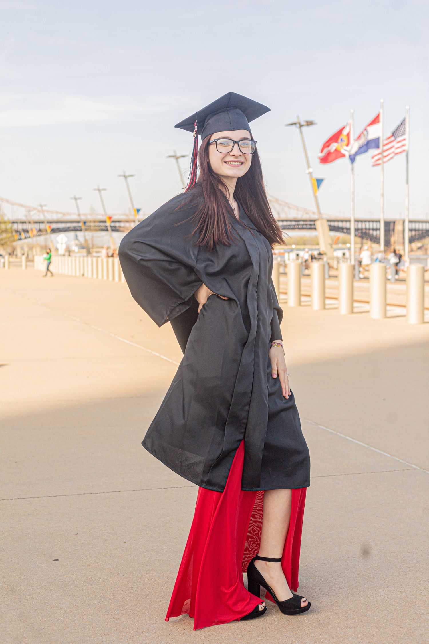Young woman in black graduation cap and gown with red dress poses outdoors near international flags on a sunny day.