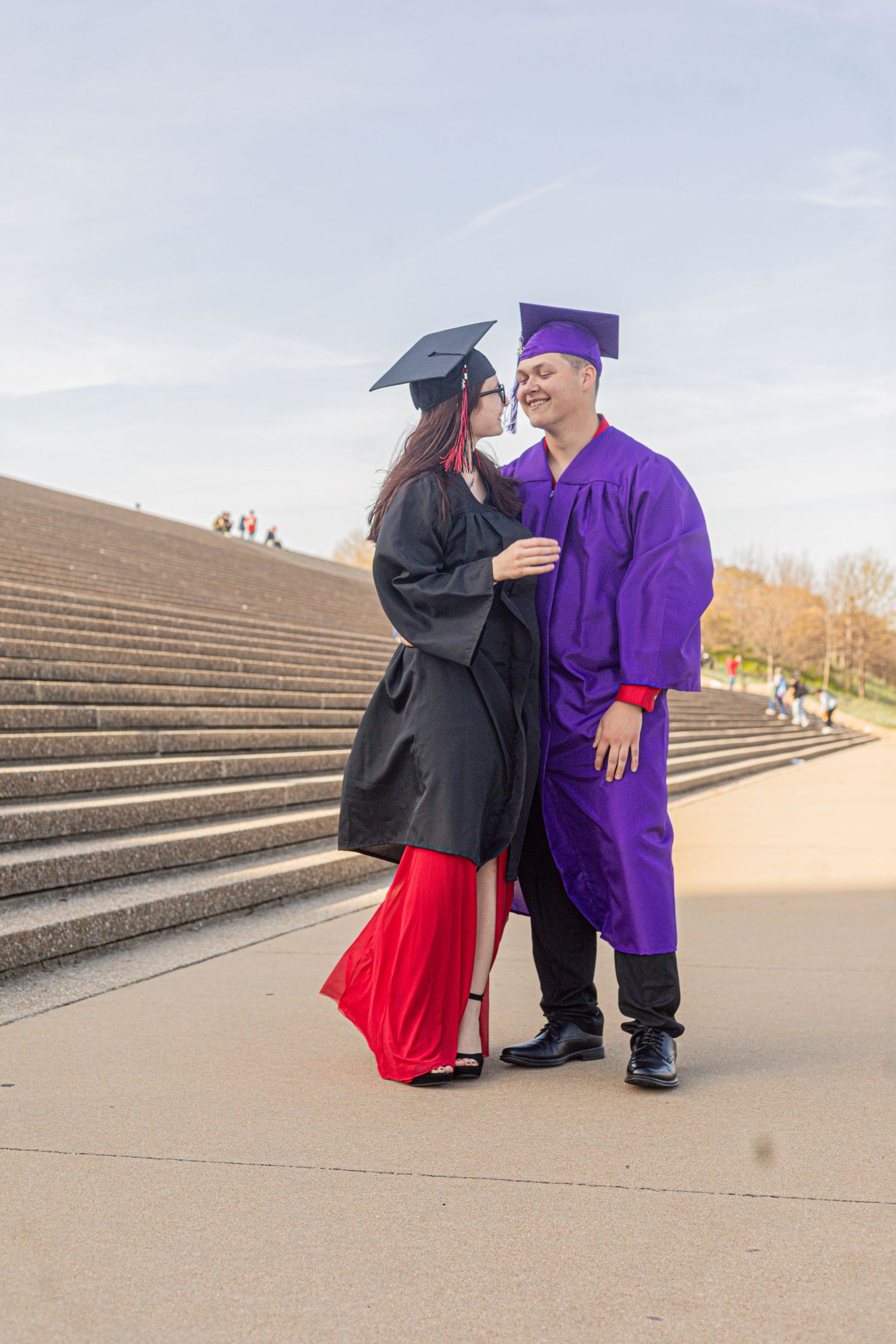 Two graduates in black and purple caps and gowns share a romantic moment on outdoor steps.