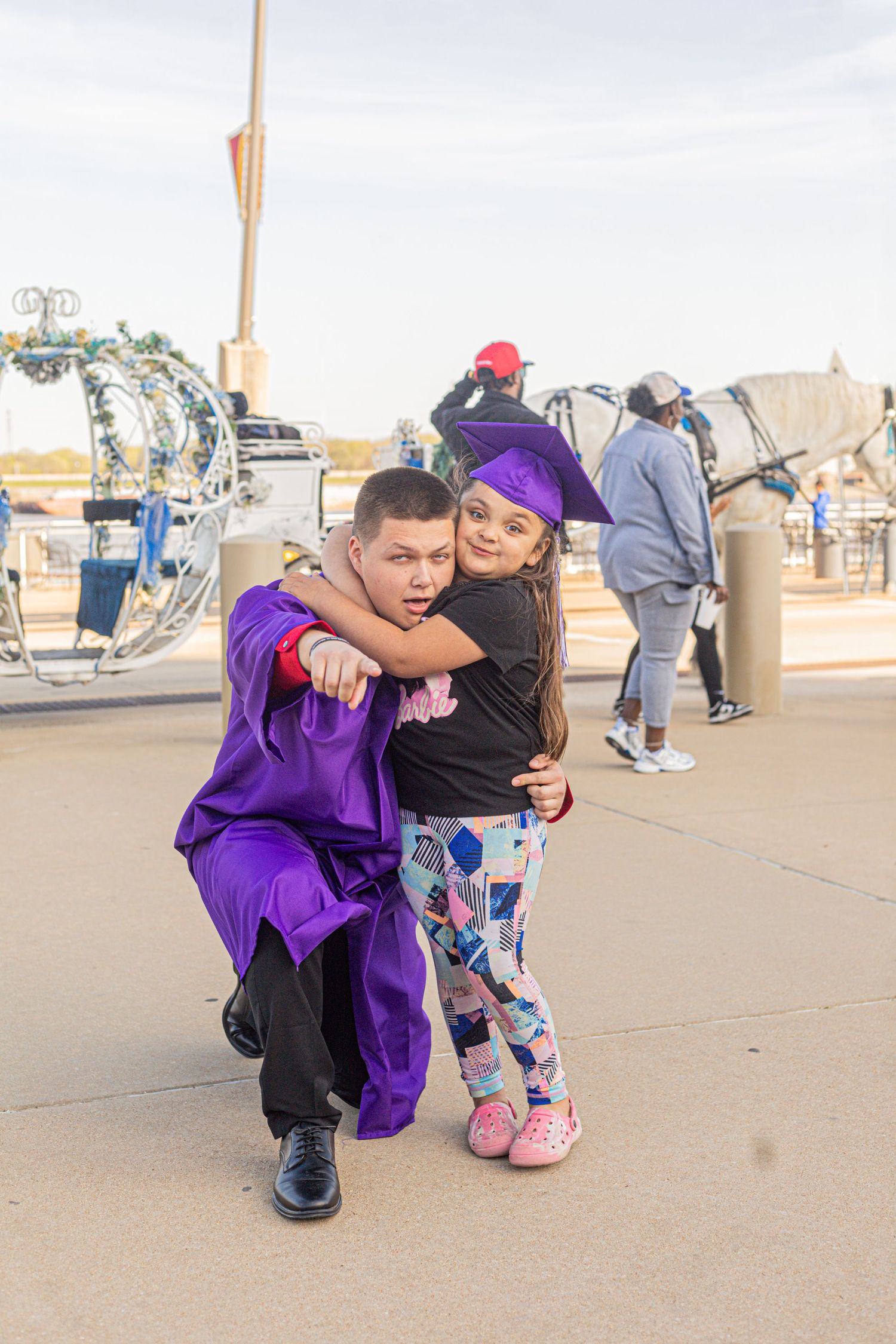 Graduate in purple cap and gown hugs young girl in colorful leggings outdoors near horse-drawn carriage.