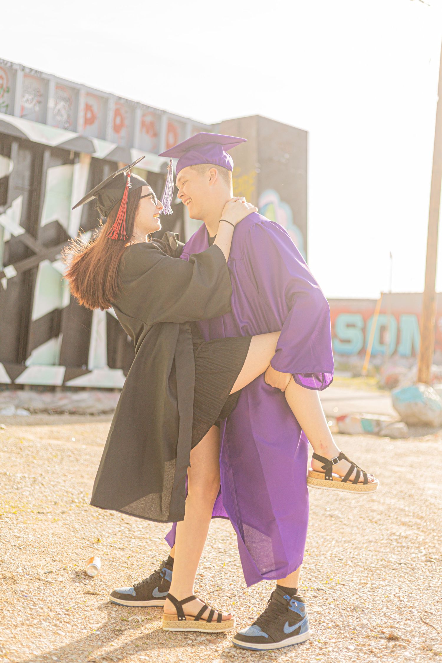 Two graduates in black and purple caps and gowns embrace outdoors, celebrating their achievement together.