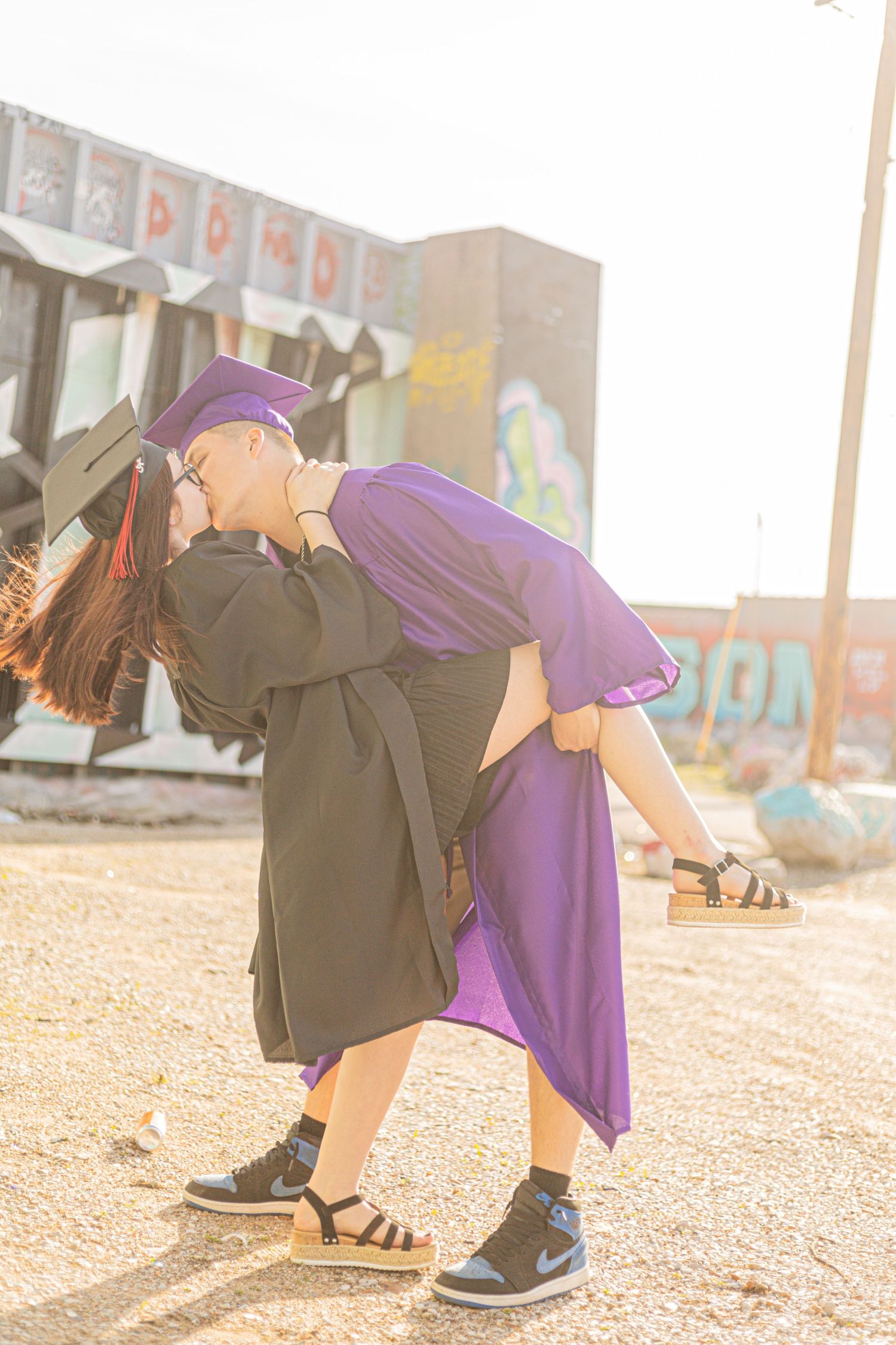Graduate in black cap and gown dips partner in purple dress for a romantic kiss outdoors near graffiti wall.