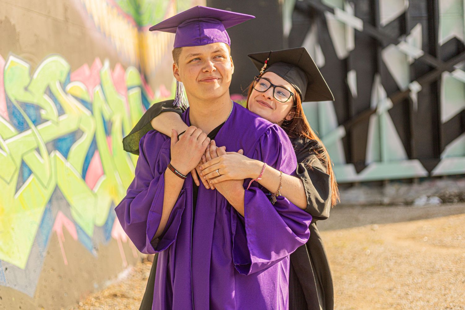 Two graduates in purple and black caps and gowns embrace warmly in front of a colorful graffiti wall.