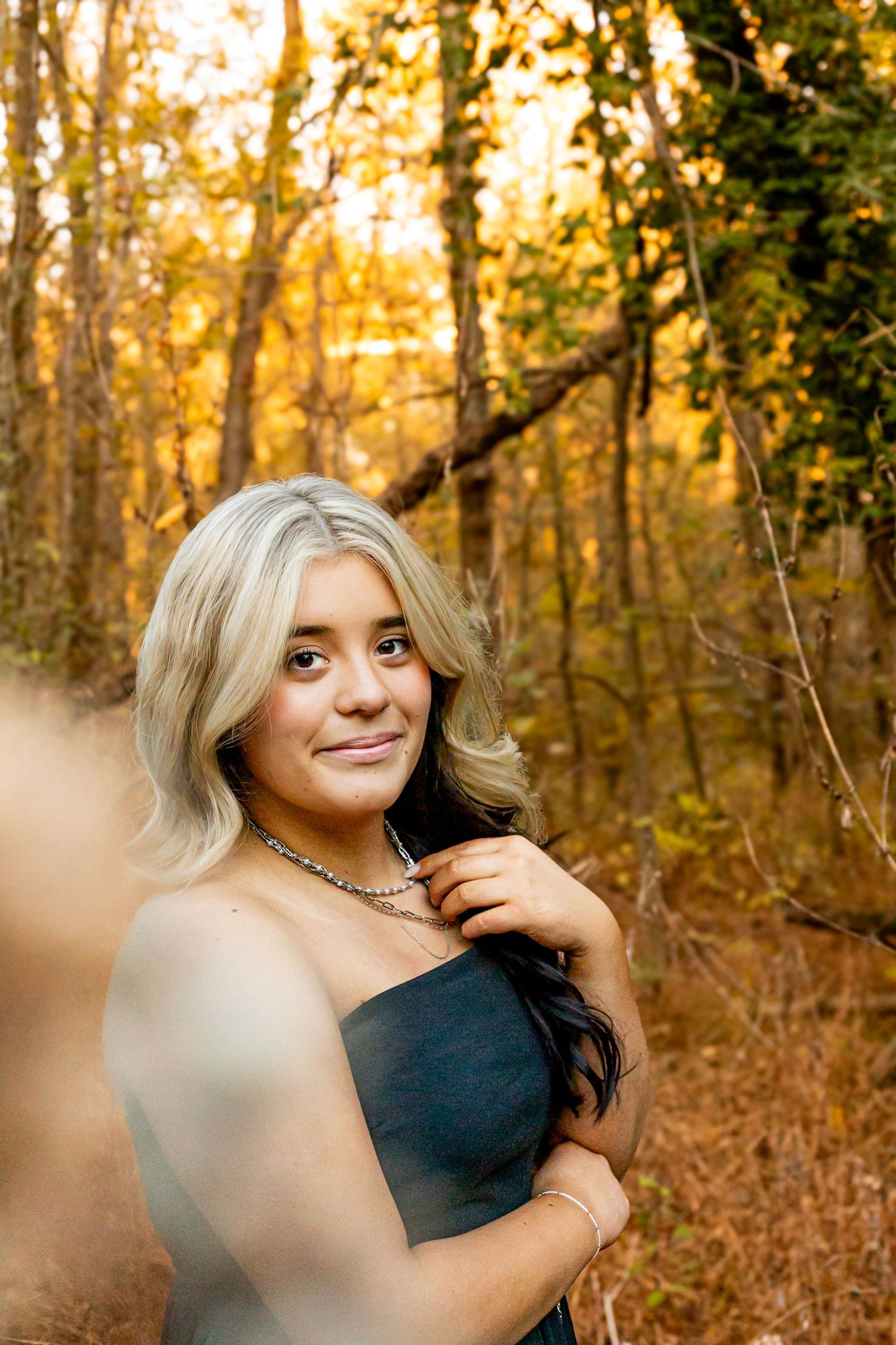 Young woman with blonde and dark hair in a black dress poses in an autumn forest with golden foliage.