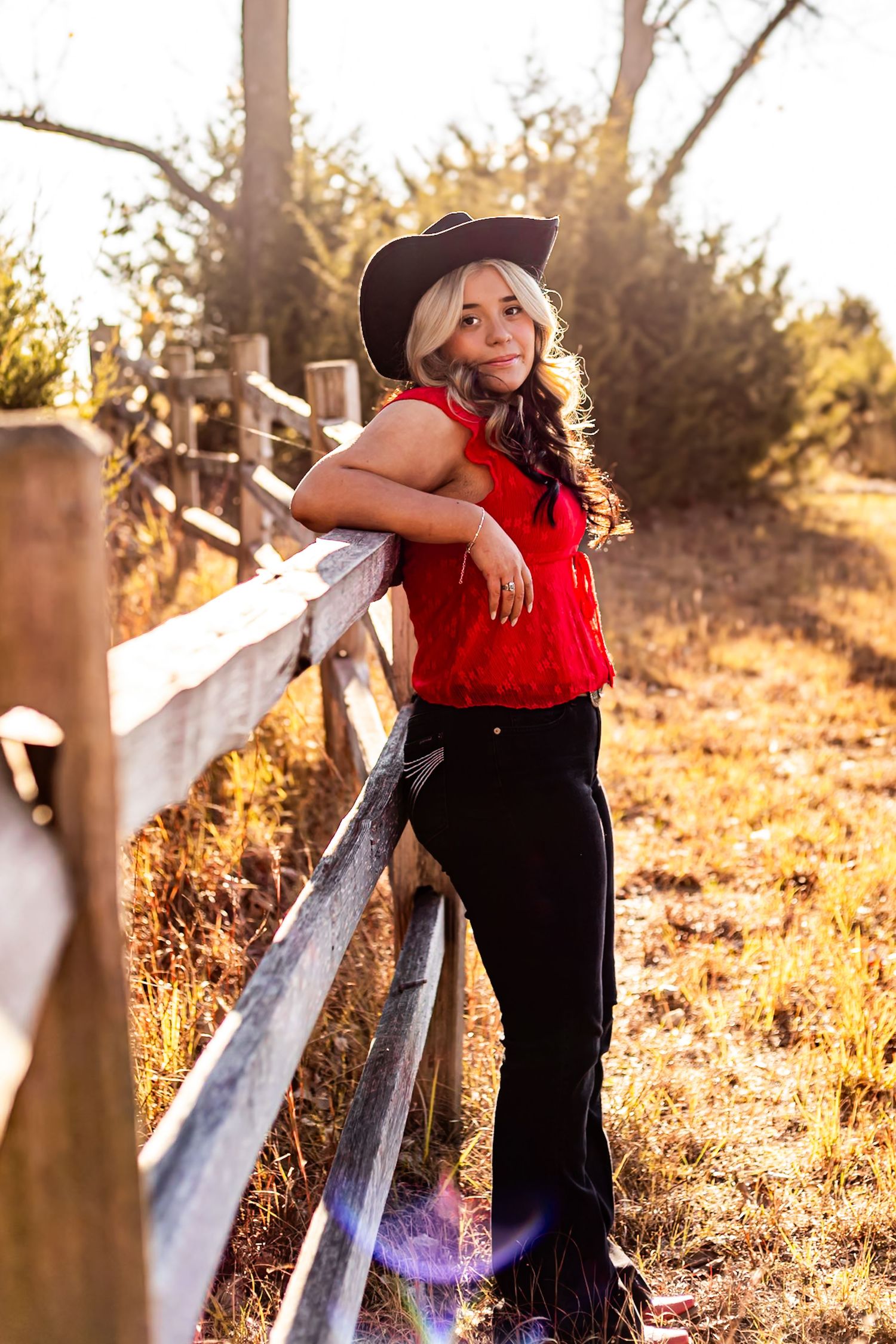 Blonde woman in red top, black cowboy hat and pants leaning on rustic wooden fence in golden sunlight.